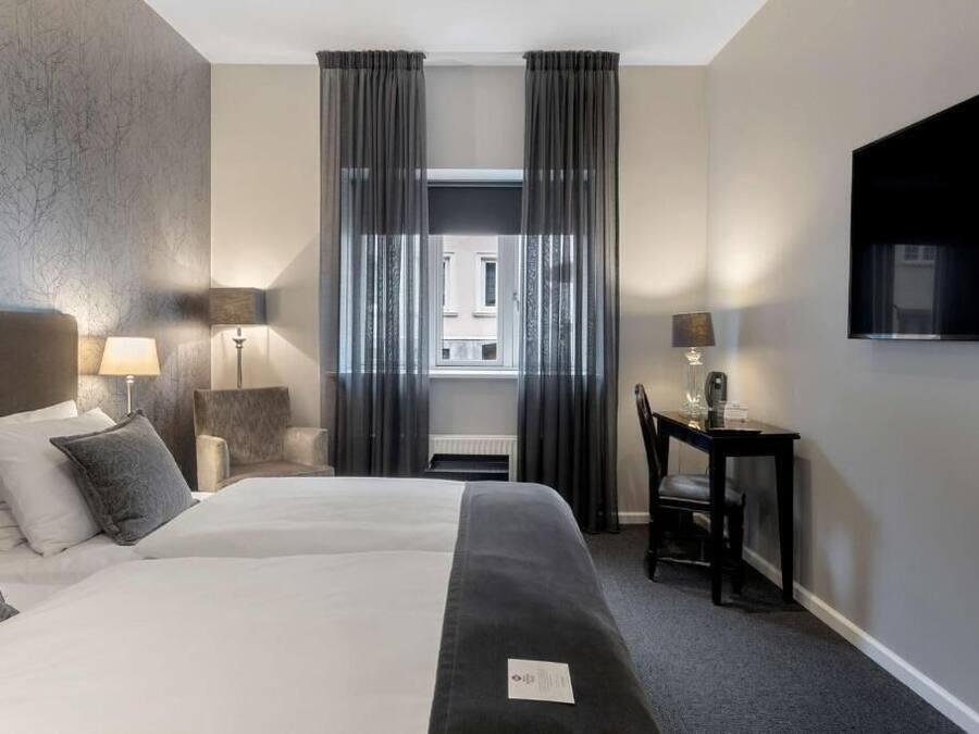 A hotel room with a large bed, two bedside lamps, a desk with a chair, a flat-screen TV, and a view of the street through the window.
