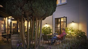Un hombre leyendo en un patio al aire libre, con una mesa, sillas y un árbol grande.