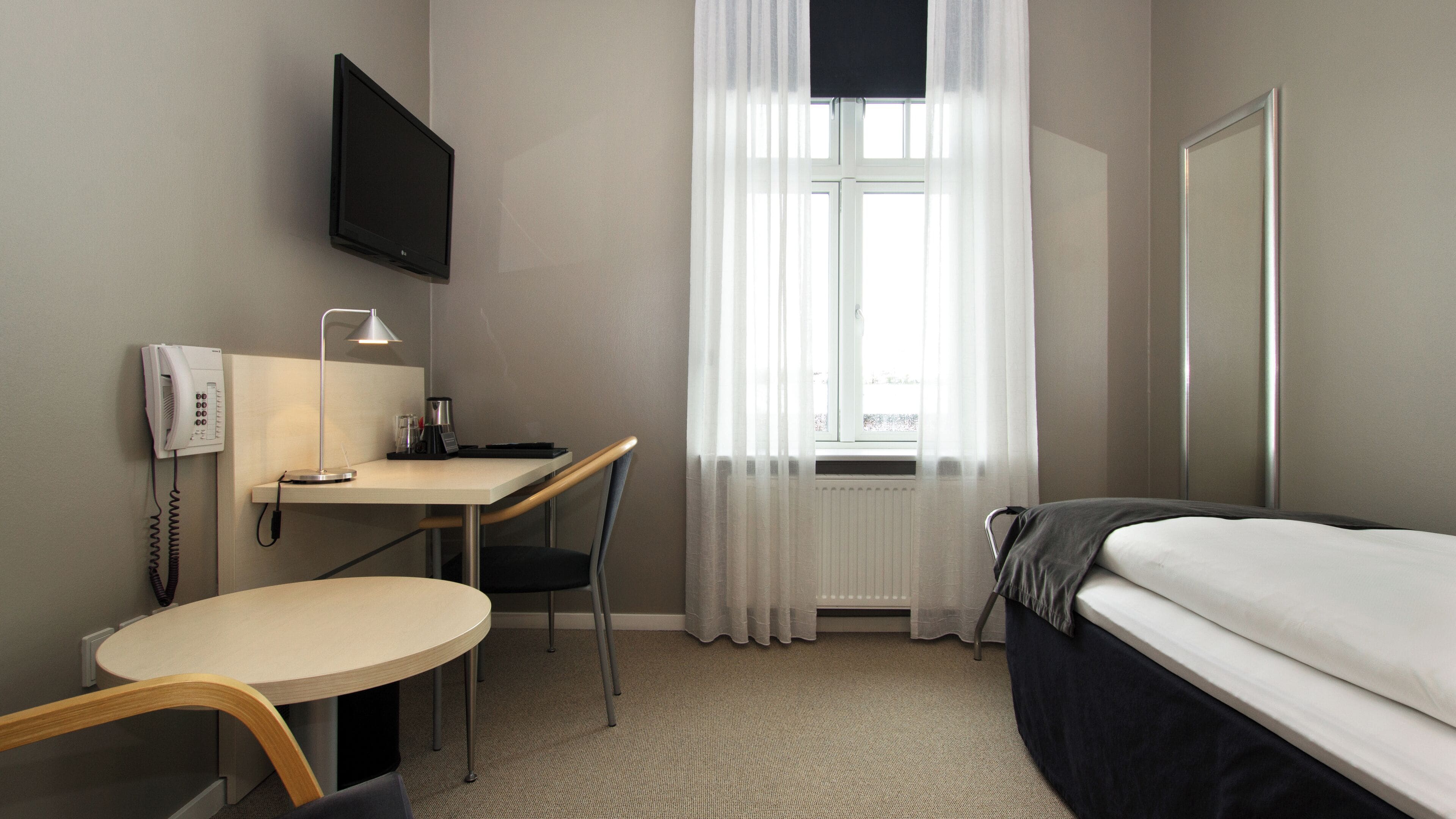 Una habitación de hotel con cama, escritorio con silla, televisor montado en la pared y ventana con cortinas translúcidas.