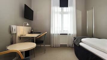 Una habitación de hotel con cama, escritorio con silla, televisor montado en la pared y ventana con cortinas translúcidas.