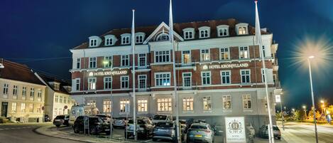 A multi-story hotel with a classic architectural style, featuring a prominent sign for 'HOTEL KRONJYLLAND' and a parking area with several cars.