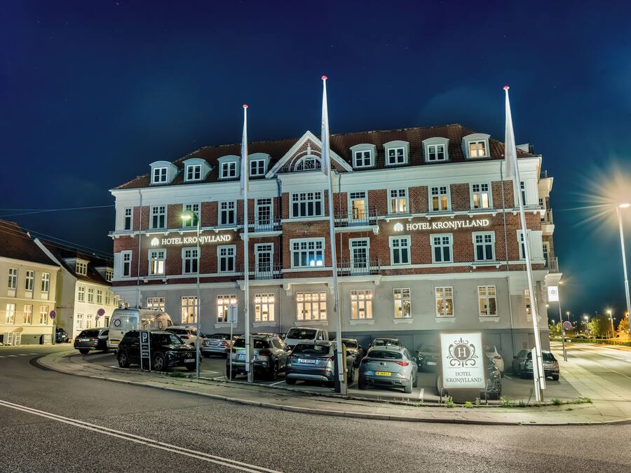 A multi-story hotel with a classic architectural style, featuring a prominent sign for 'HOTEL KRONJYLLAND' and a parking area with several cars.