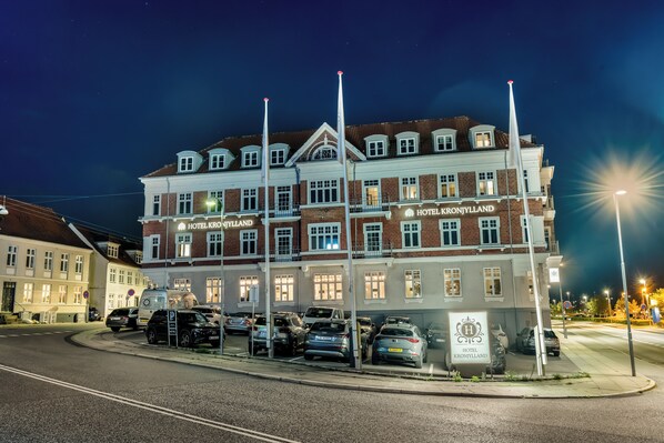 Un hotel de varios pisos con un estilo arquitectónico clásico, que presenta un letrero llamativo que dice 'HOTEL KRONJYLLAND' y un área de estacionamiento con varios coches.