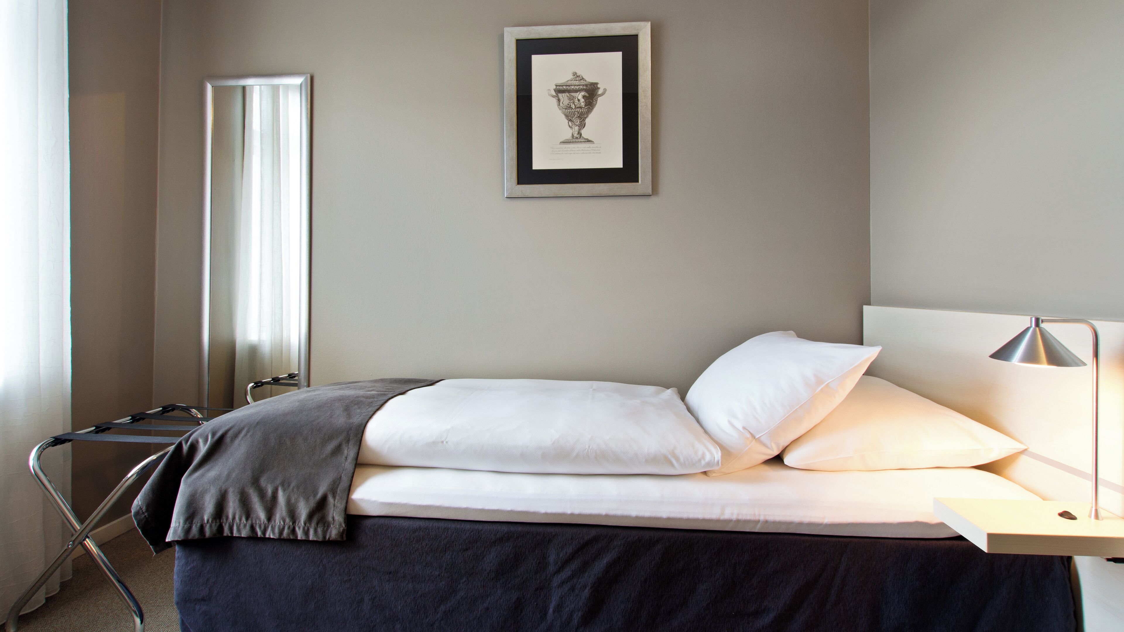 A neatly made bed with white linens, a gray throw blanket, and a bedside table with a lamp. A framed picture hangs on the wall above the bed.