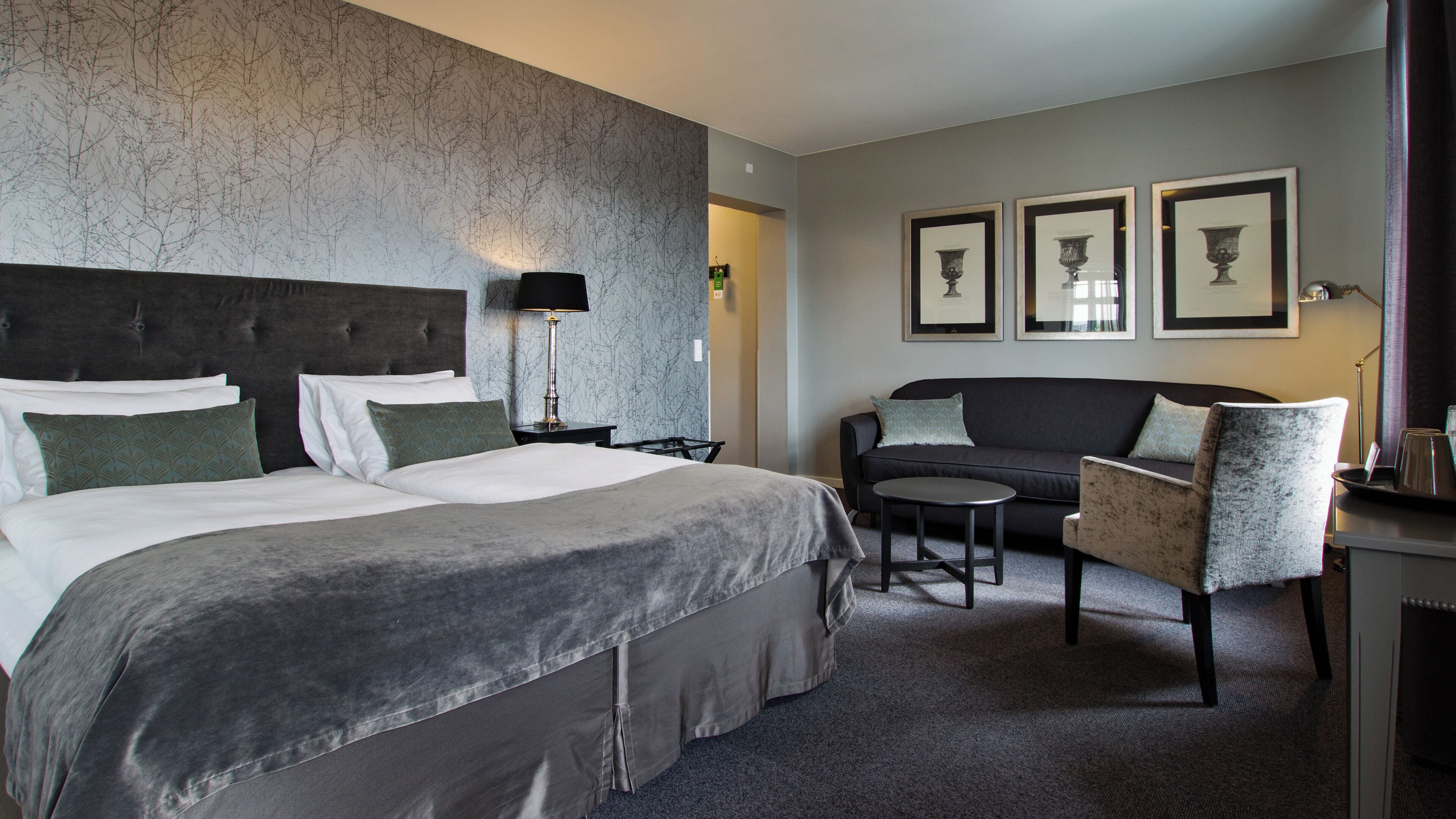 Habitación de hotel con una cama grande, un sofá, una mesita y una silla. En la pared hay cuadros enmarcados.