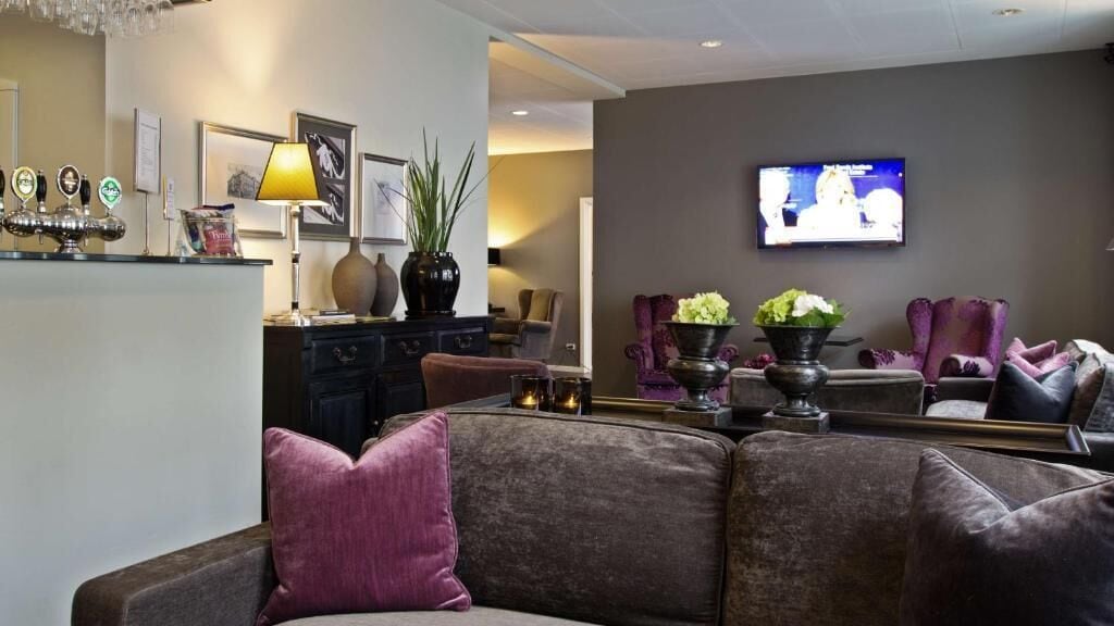 A modern living room with a large flat-screen TV mounted on the wall, a dark-colored sofa with a purple pillow, a coffee table with candles, and a bar area with beer taps.