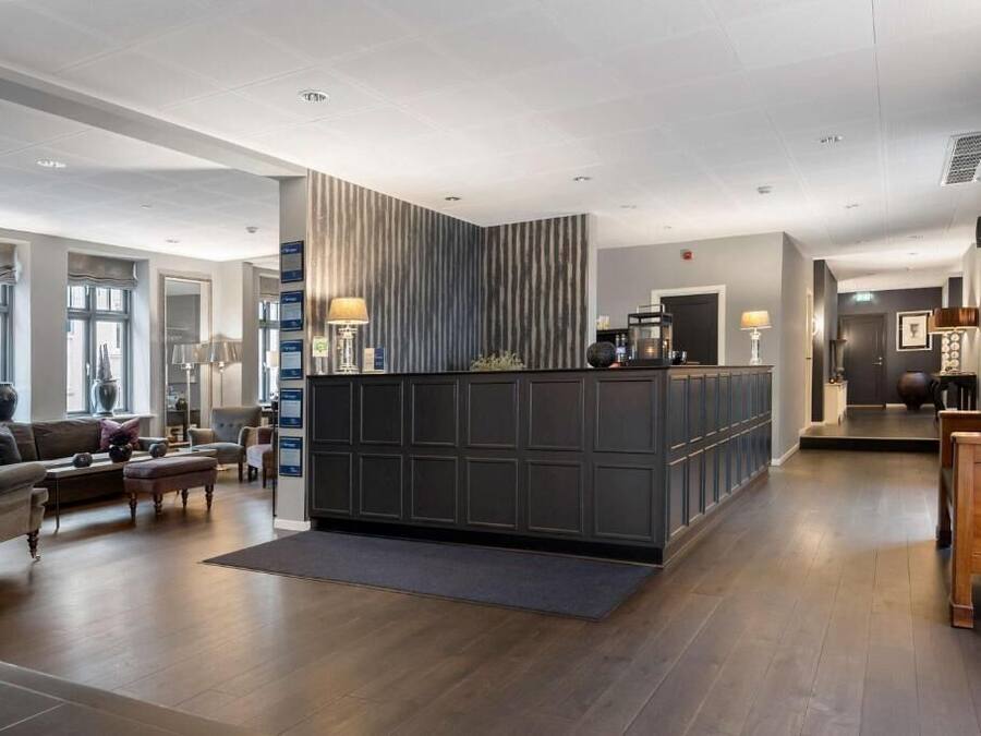 Un vestíbulo de hotel moderno con un mostrador de recepción de madera oscura, un área de descanso con un sofá y sillones, y un gran ventanal que proporciona luz natural.