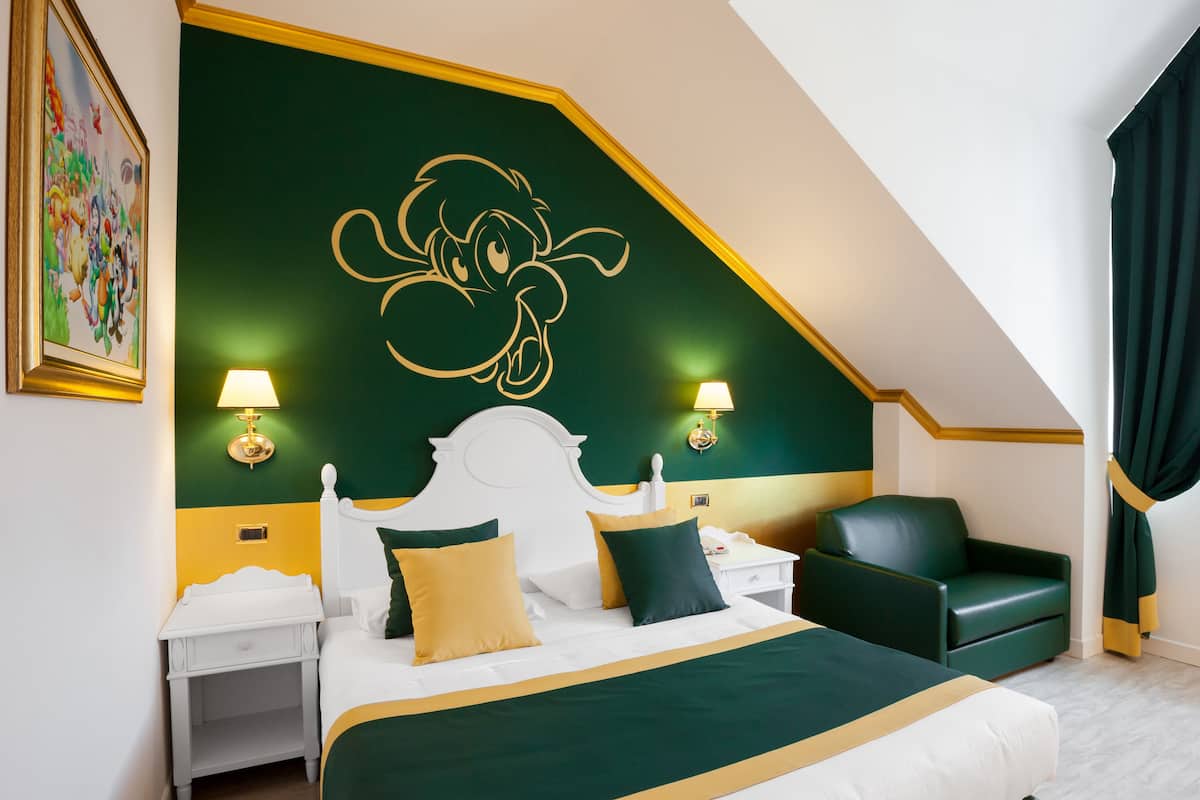 Une chambre d’hôtel avec un grand lit, deux tables de chevet, un fauteuil vert et un tableau au mur.