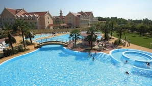 A large swimming pool with a bridge, surrounded by palm trees and a building with a red roof. - Gardaland Hotel (Castelnuovo del Garda)