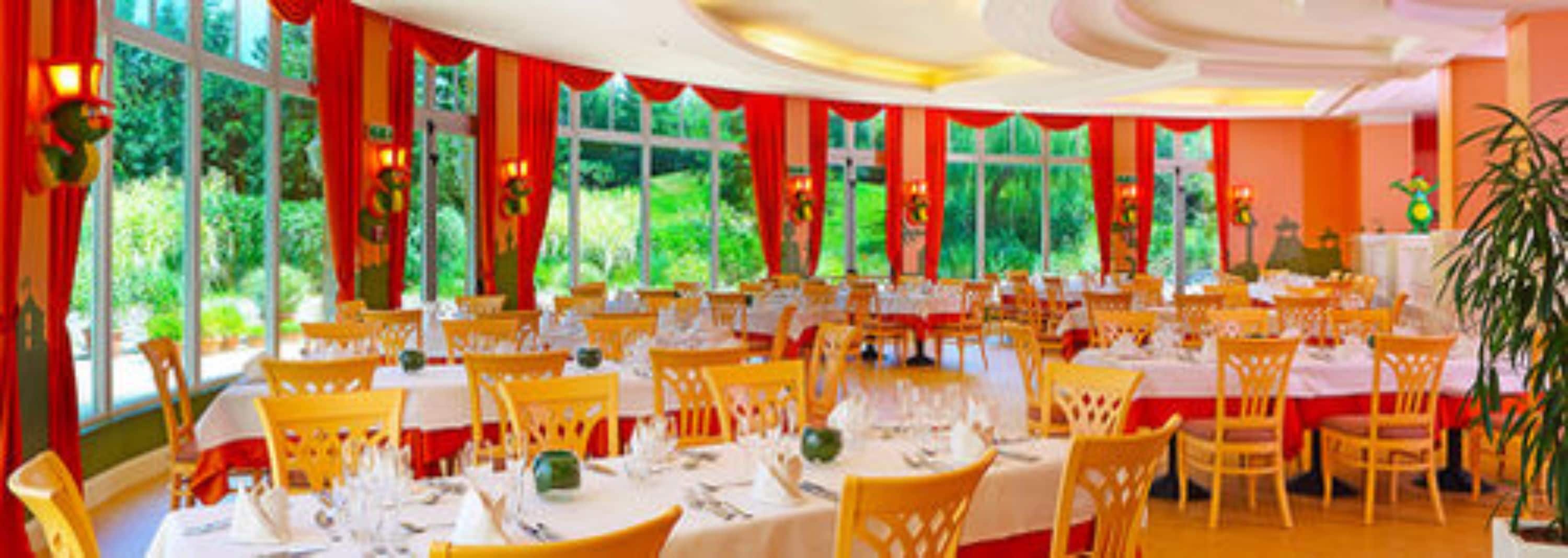 A large dining area with round tables and yellow chairs, large windows with red curtains, and a view of greenery outside.