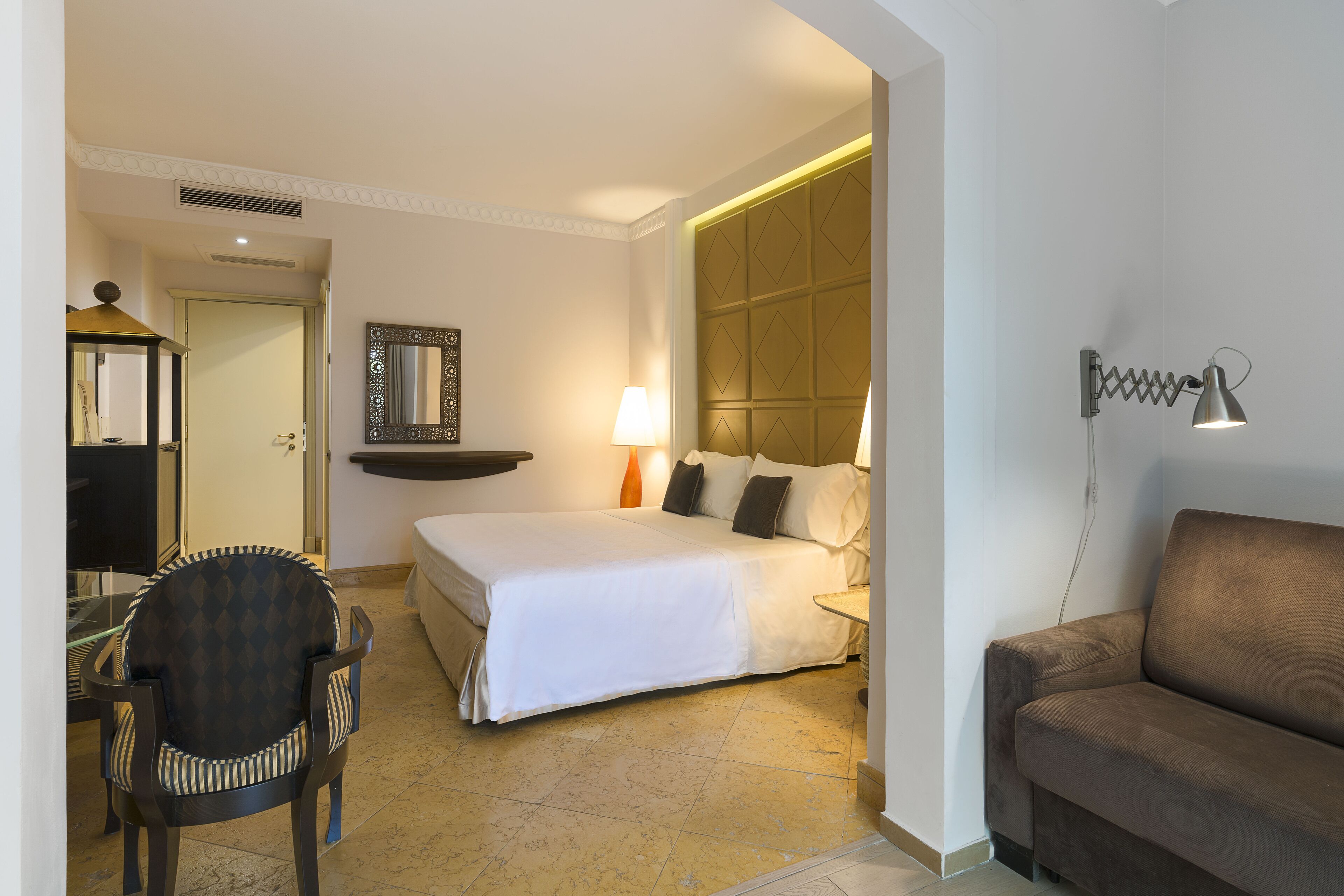 A hotel room with a bed, a sofa, a chair, a desk, a lamp, and a mirror.