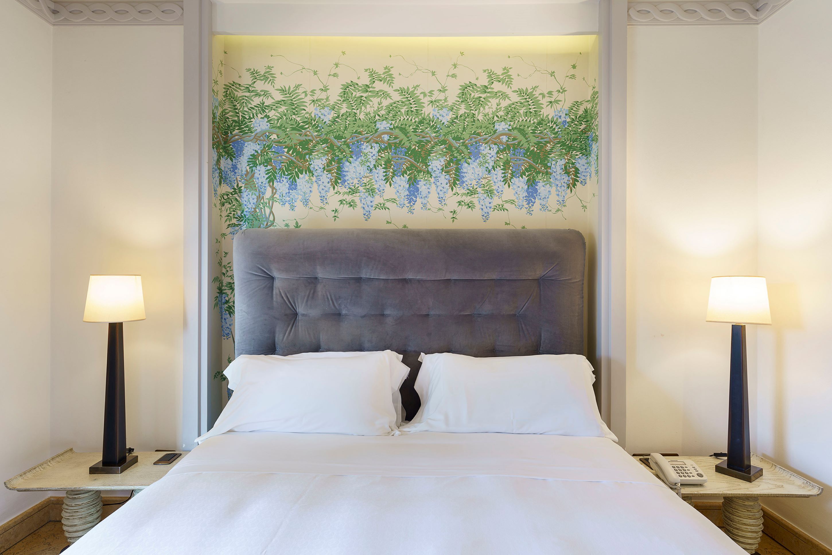 A hotel room with a large bed, two bedside tables with lamps, and a wall mural of hanging plants.