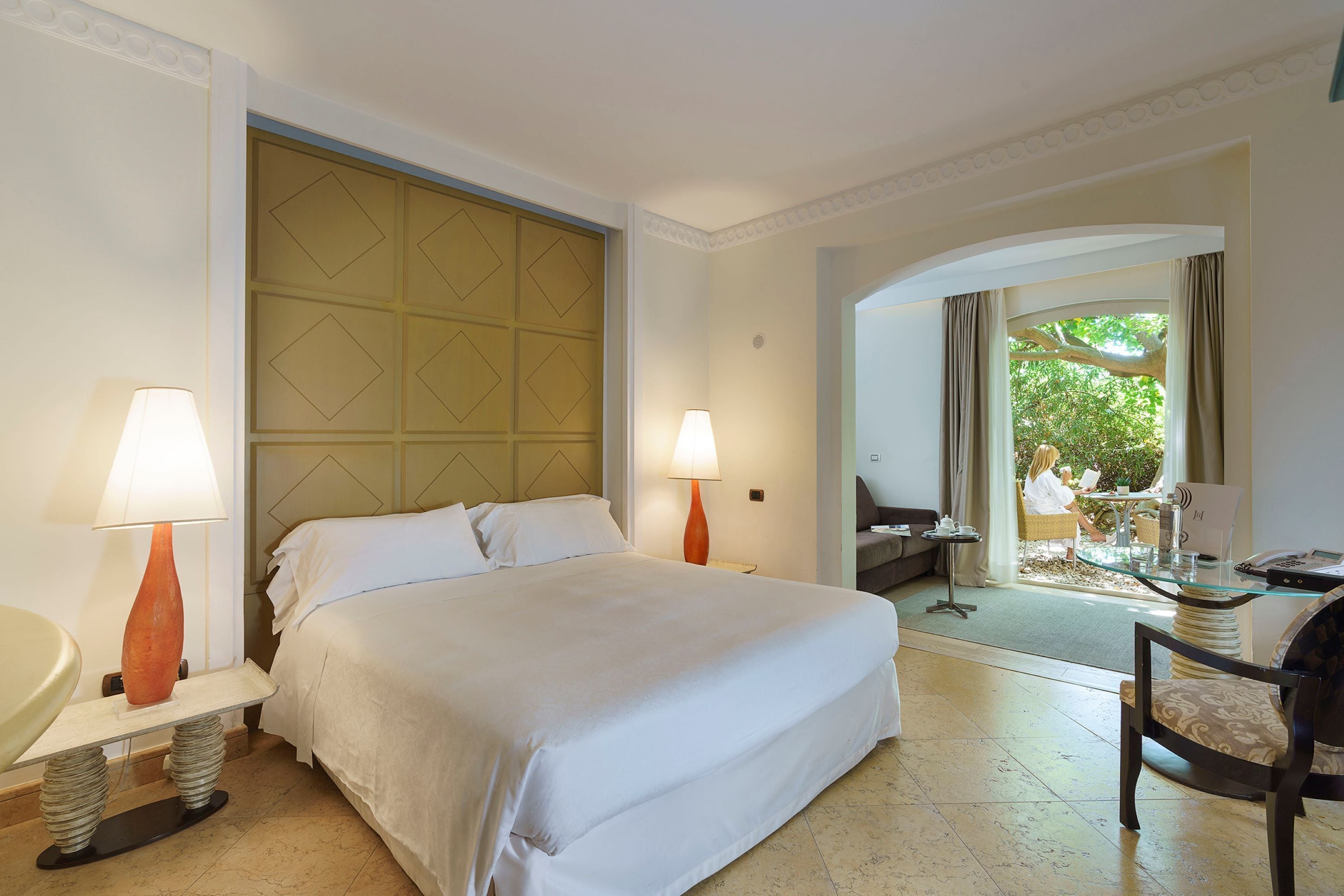 Une chambre d’hôtel avec un grand lit, deux lampes de chevet, une petite table avec une chaise, et une vue sur l’extérieur par une porte.