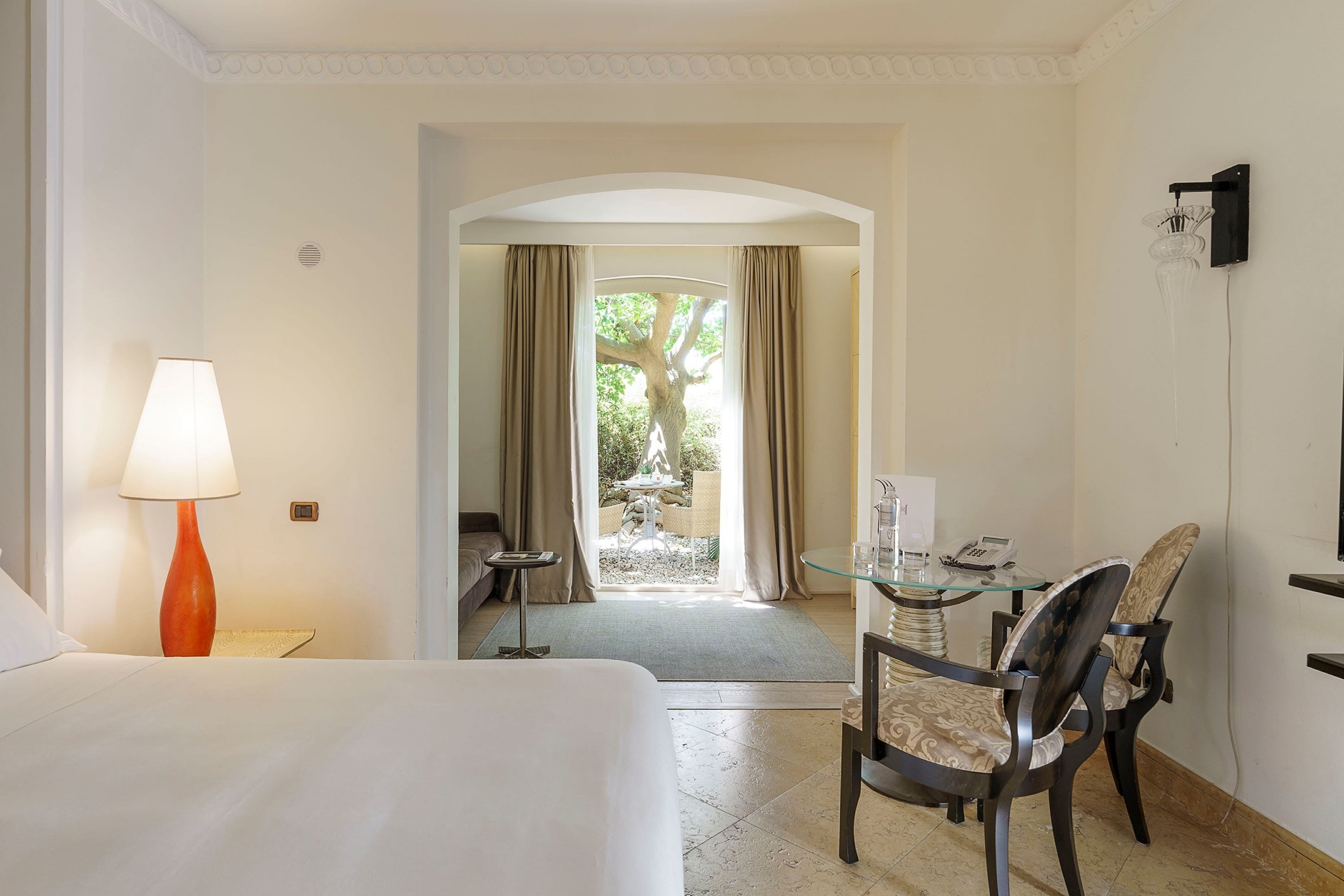 A hotel room with a bed, a table with chairs, a lamp, and a view of the outdoors.