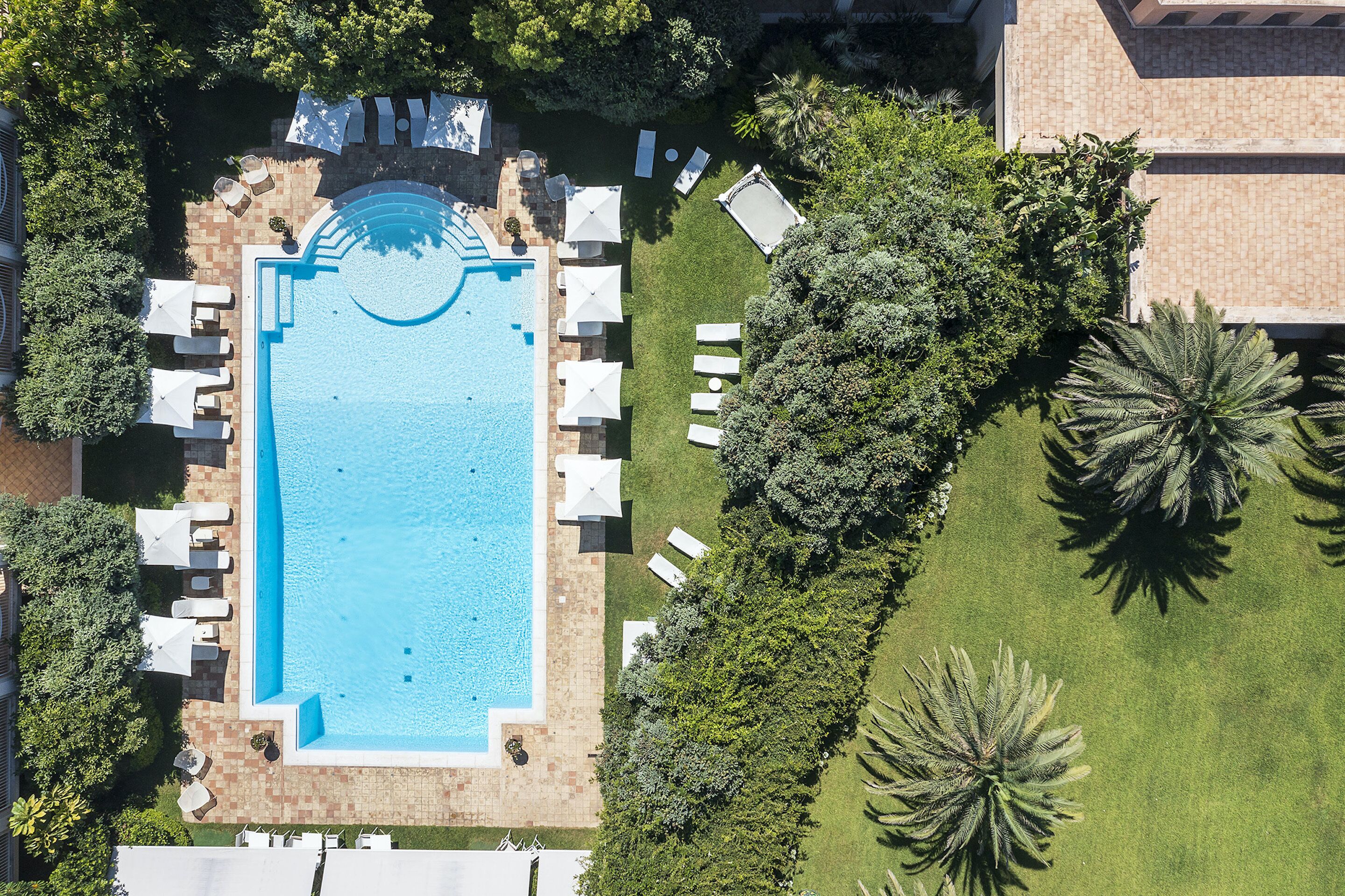 An aerial view of a swimming pool surrounded by lounge chairs and umbrellas, with lush greenery and palm trees.