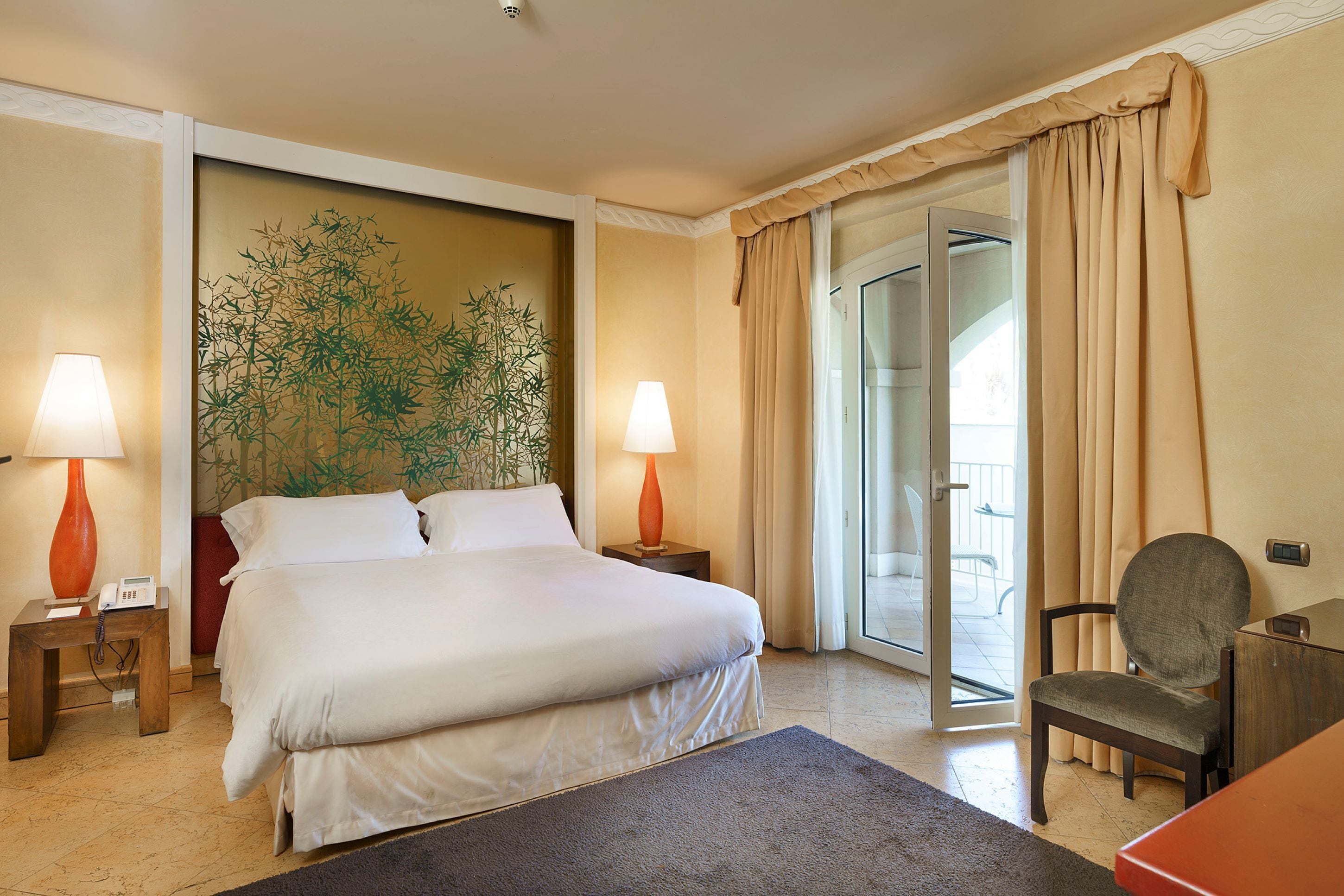 A hotel room with a large bed, two bedside tables with lamps, a chair, and a view of the outdoors through a glass door.