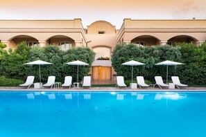 Zona de piscina con sillones blancos y sombrillas, rodeada de follaje y un edificio con arcos.