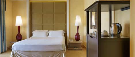 Quarto de hotel com uma cama, duas luminárias de cabeceira, uma mesa de apoio, um telefone e um armário com chaleira.