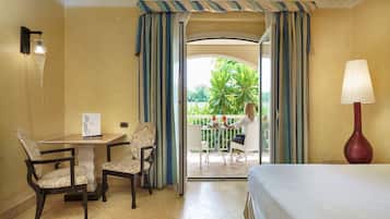 Ein Hotelzimmer mit Bett, zwei Stühlen, einem Tisch, einer Lampe und Blick auf einen Balkon, auf dem eine Person sitzt.
