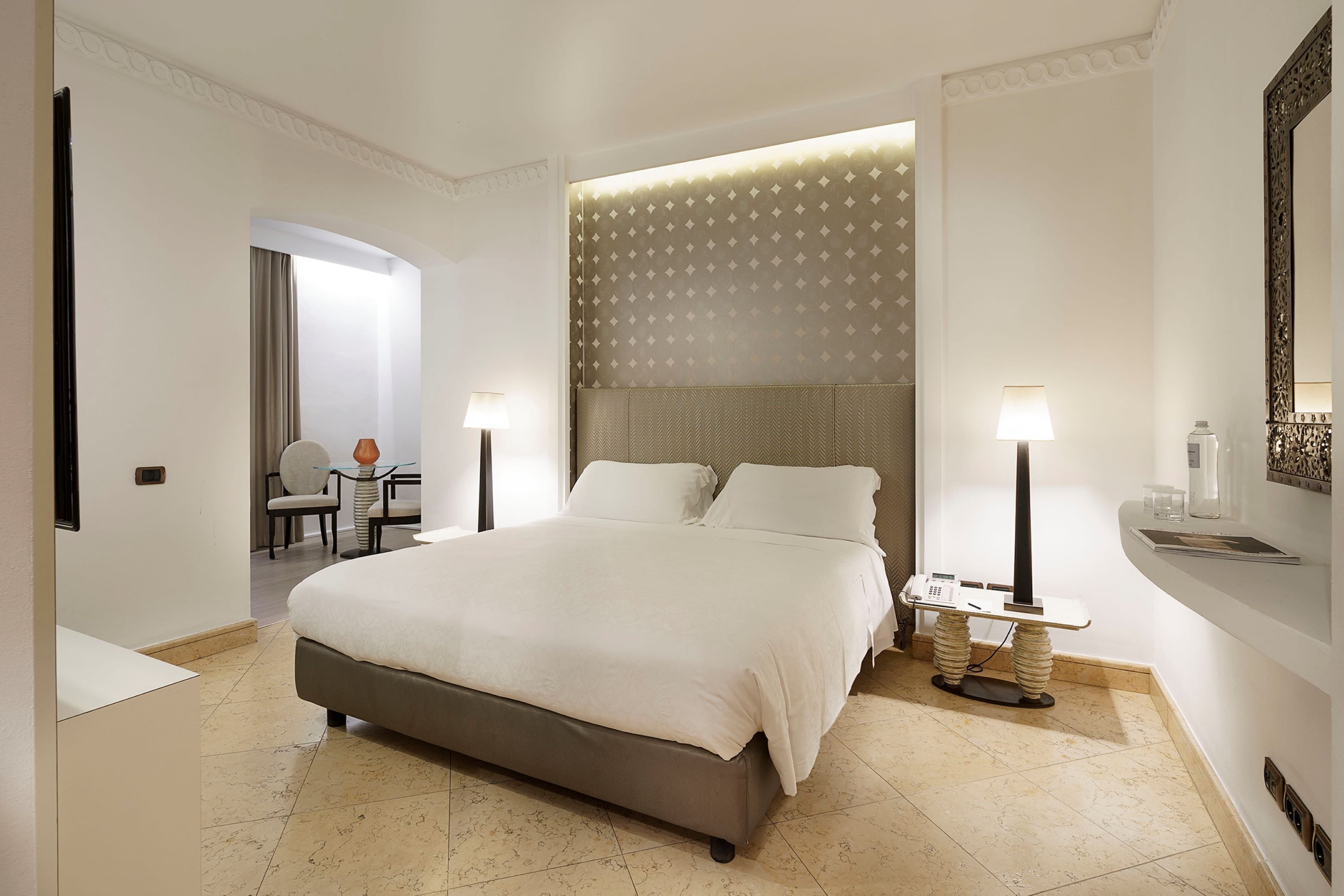 A modern hotel room with a large bed, two bedside tables with lamps, a small seating area with a chair and table, and a decorative headboard.