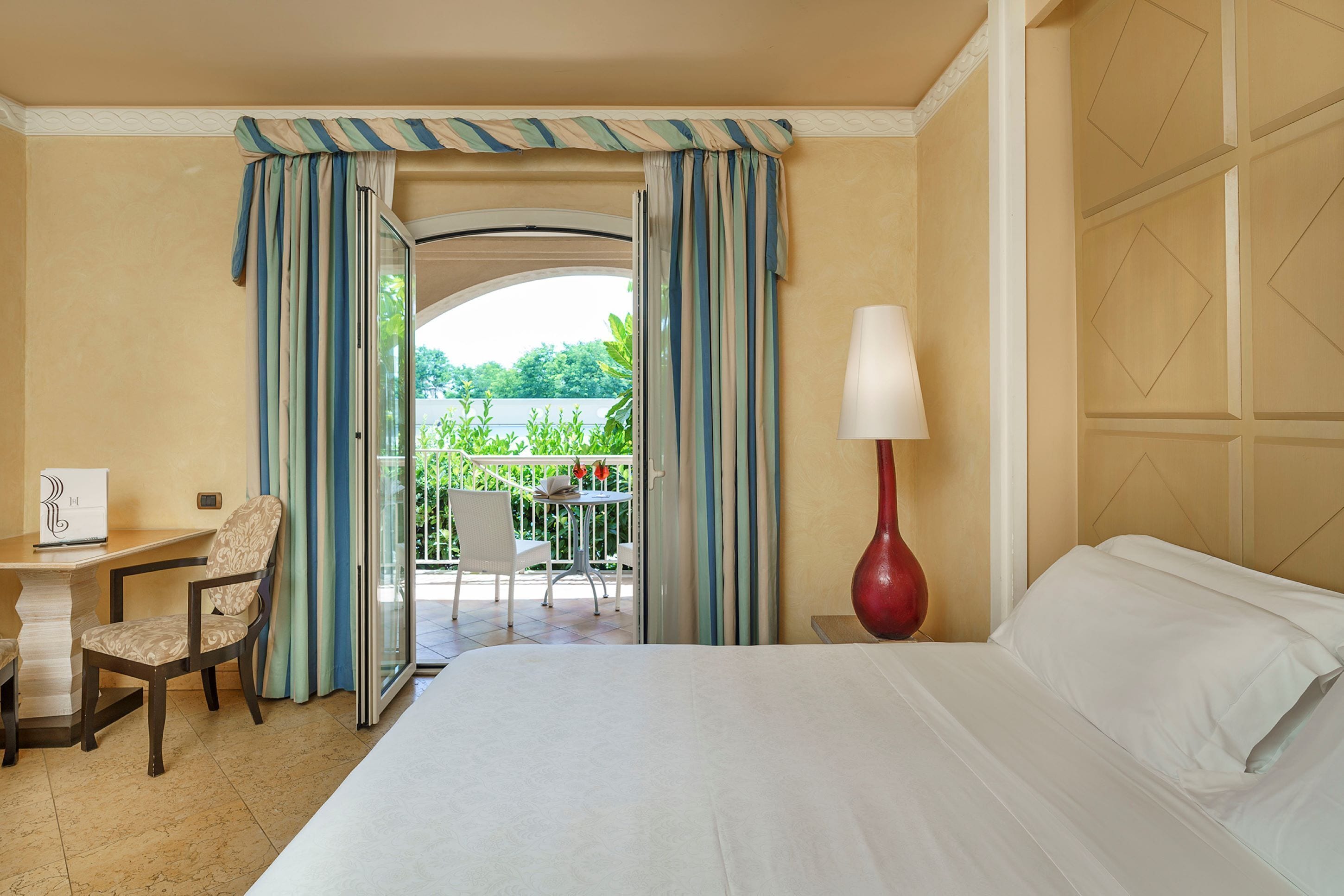 Habitación de hotel con cama, escritorio, silla, lámpara y vistas al exterior.