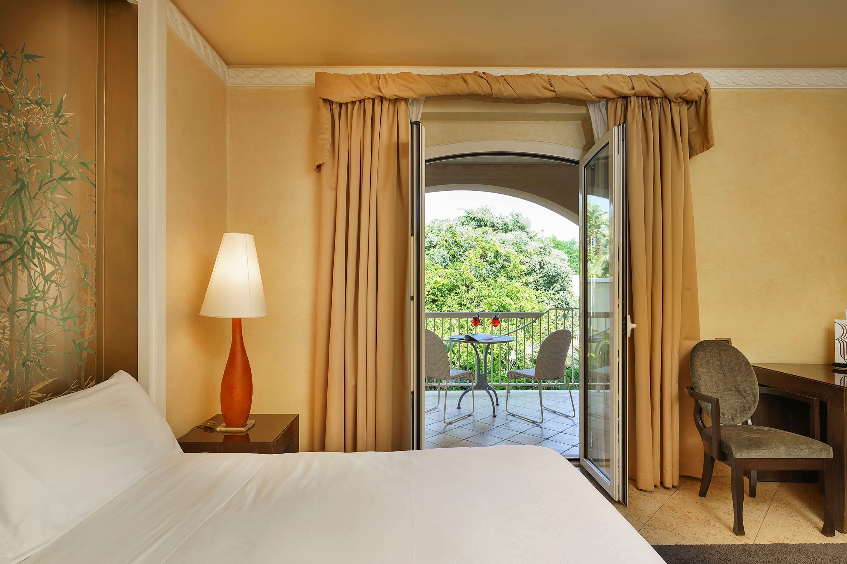 Une chambre d’hôtel comprenant un lit, un bureau, une chaise, une lampe et un balcon avec une table et des chaises.