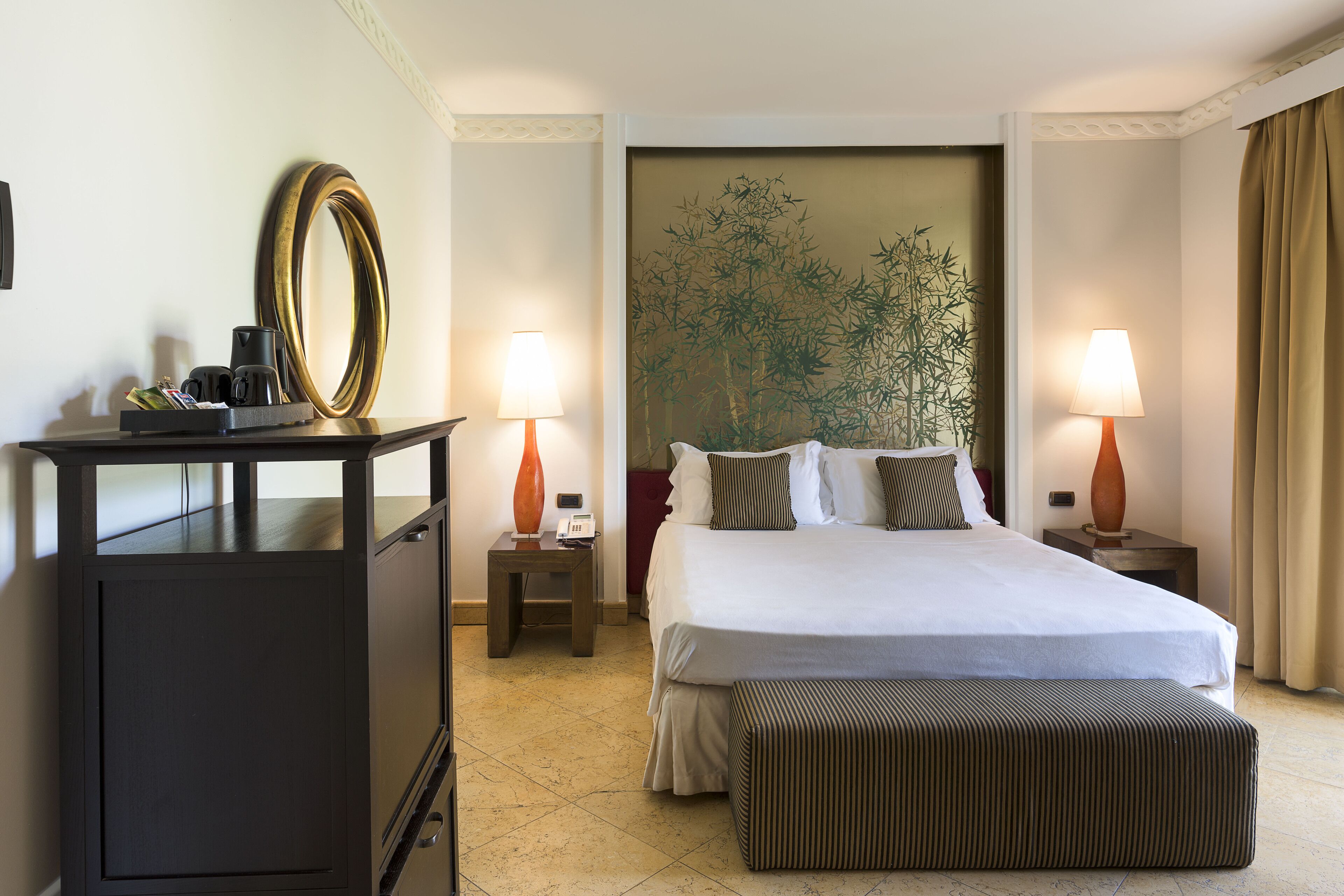 A hotel room with a bed, bedside tables, a desk with a mirror, and a large wall painting.
