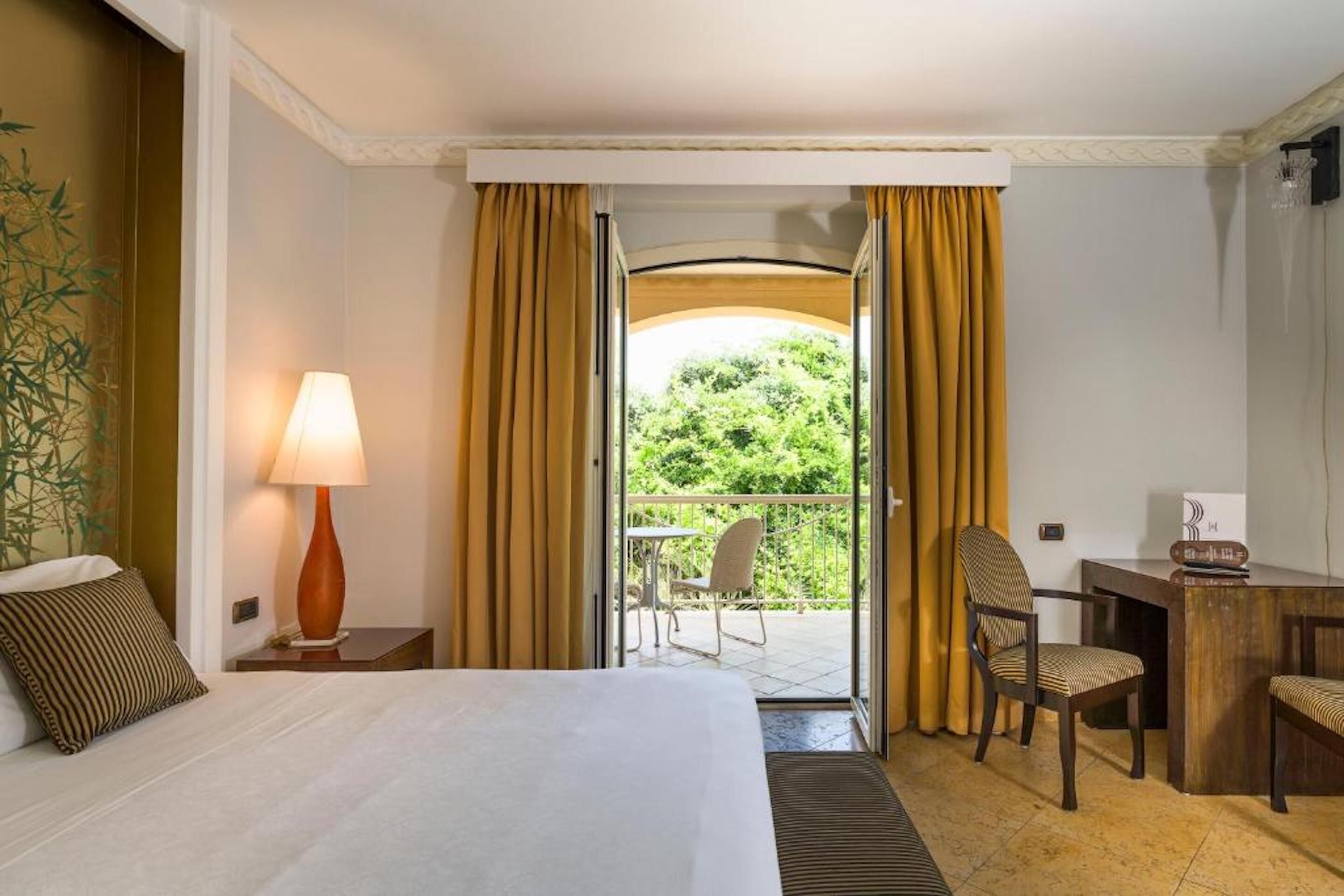 Habitación de hotel con cama, escritorio, silla, lámpara y vista a zonas verdes a través de una puerta abierta.