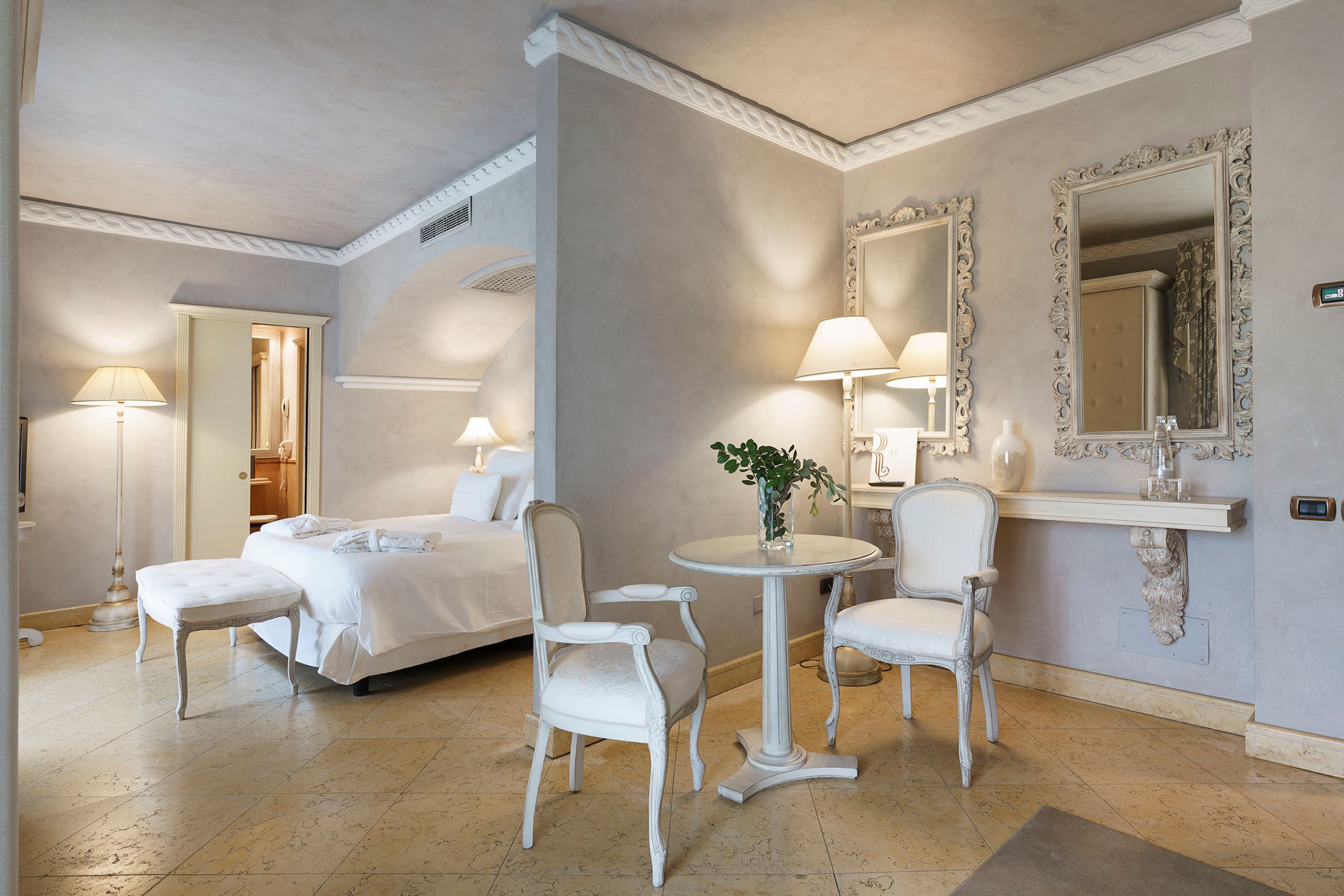 Habitación de hotel con cama, dos sillas, una mesa redonda con planta y un espejo con marcos ornamentados.