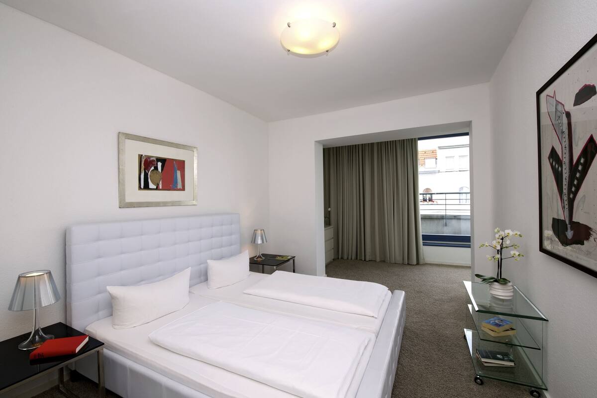 Una habitación de hotel con una cama grande, una mesita de noche con una lámpara, una pequeña mesita auxiliar, un aparador de vidrio con libros, y un cuadro en la pared.