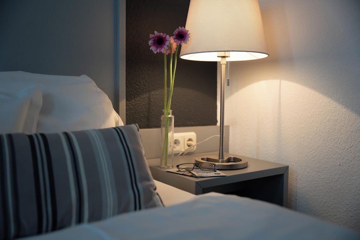 A neatly made bed with a striped pillow, a bedside table with a lamp, and a vase with flowers.