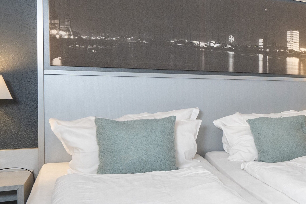 A hotel room with two beds, a nightstand with a lamp, and a cityscape view on the wall.