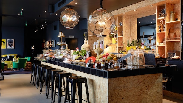 A bar area with stools, a counter with food and drink items, and hanging pendant lights. - Hotel Medo Köln (Cologne)