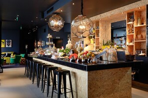 A bar area with stools, a counter with food and drink items, and hanging pendant lights. - Hotel Medo Köln (Cologne)