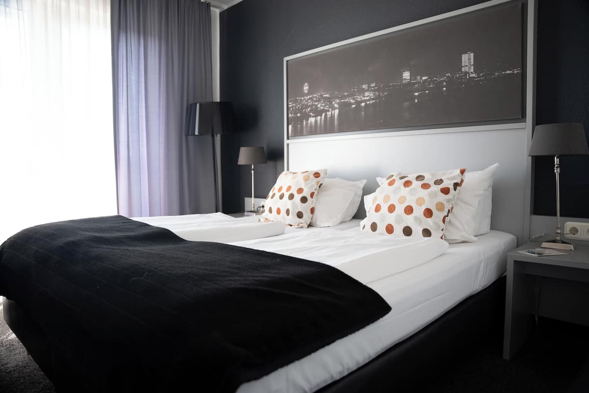 A hotel room with a large bed, a cityscape view, and a desk with a lamp.