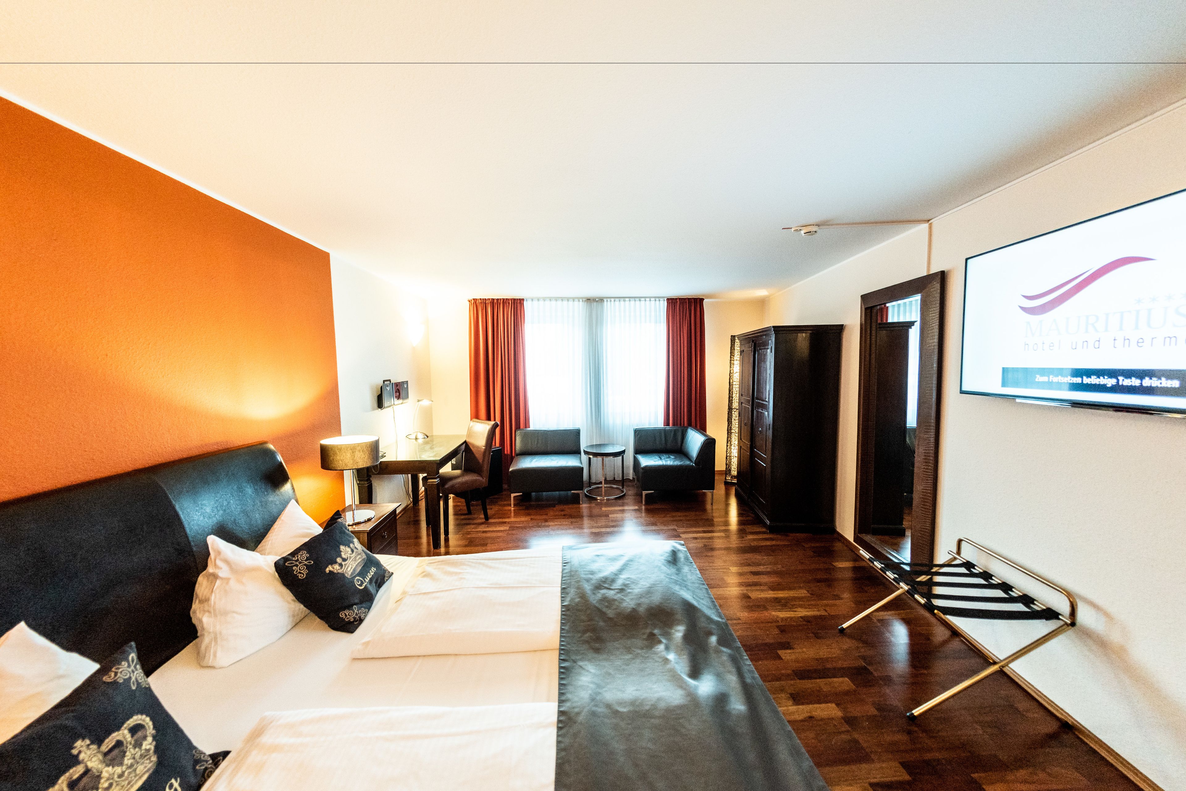 A hotel room with a bed, a desk, a chair, a sofa, and a screen displaying the hotel name.