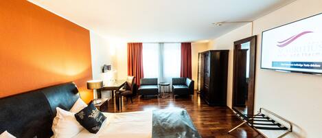 A hotel room with a bed, a desk, a chair, a sofa, and a screen displaying the hotel name.