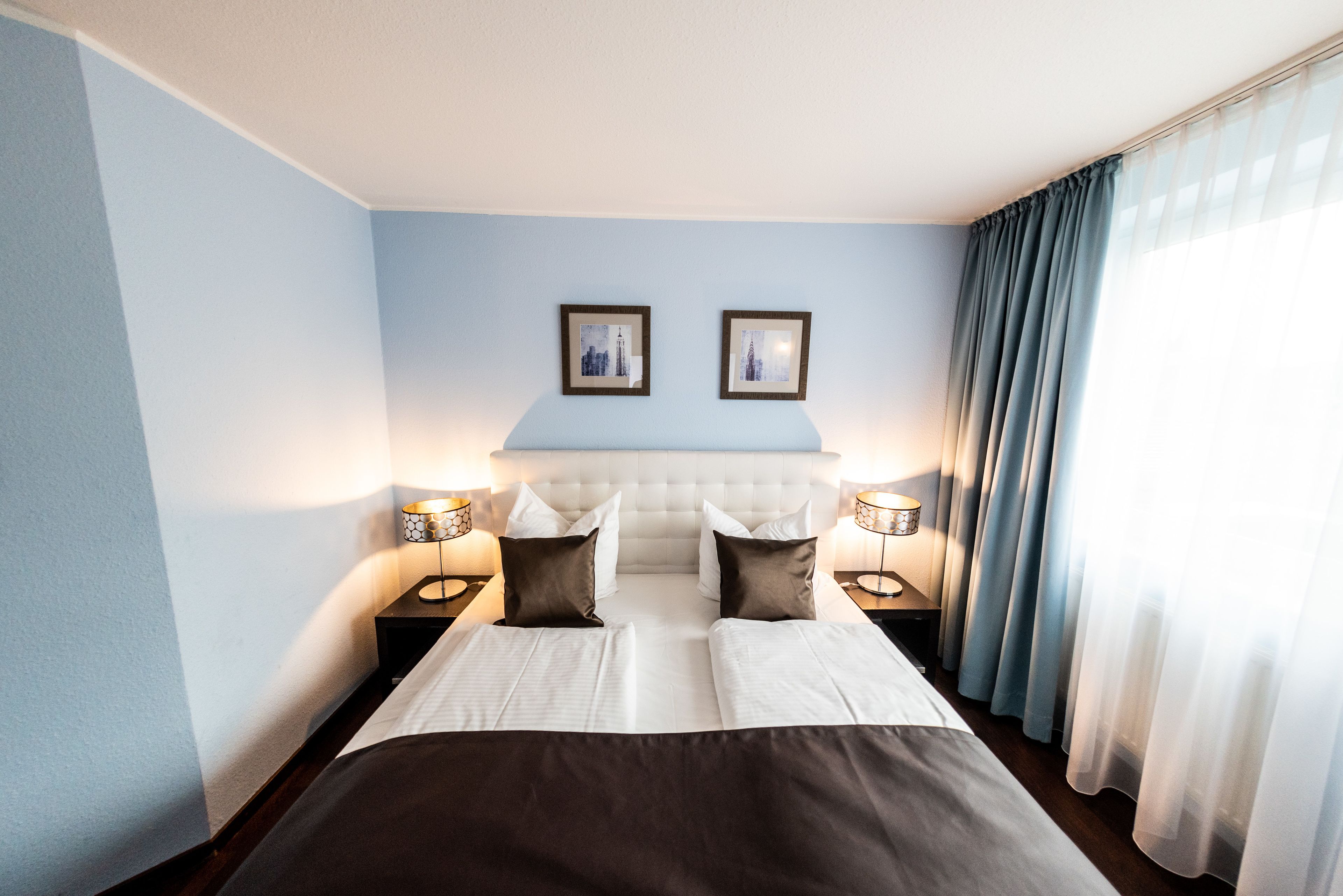 A hotel room with a large bed, two bedside tables with lamps, a headboard, and two framed pictures on the wall.