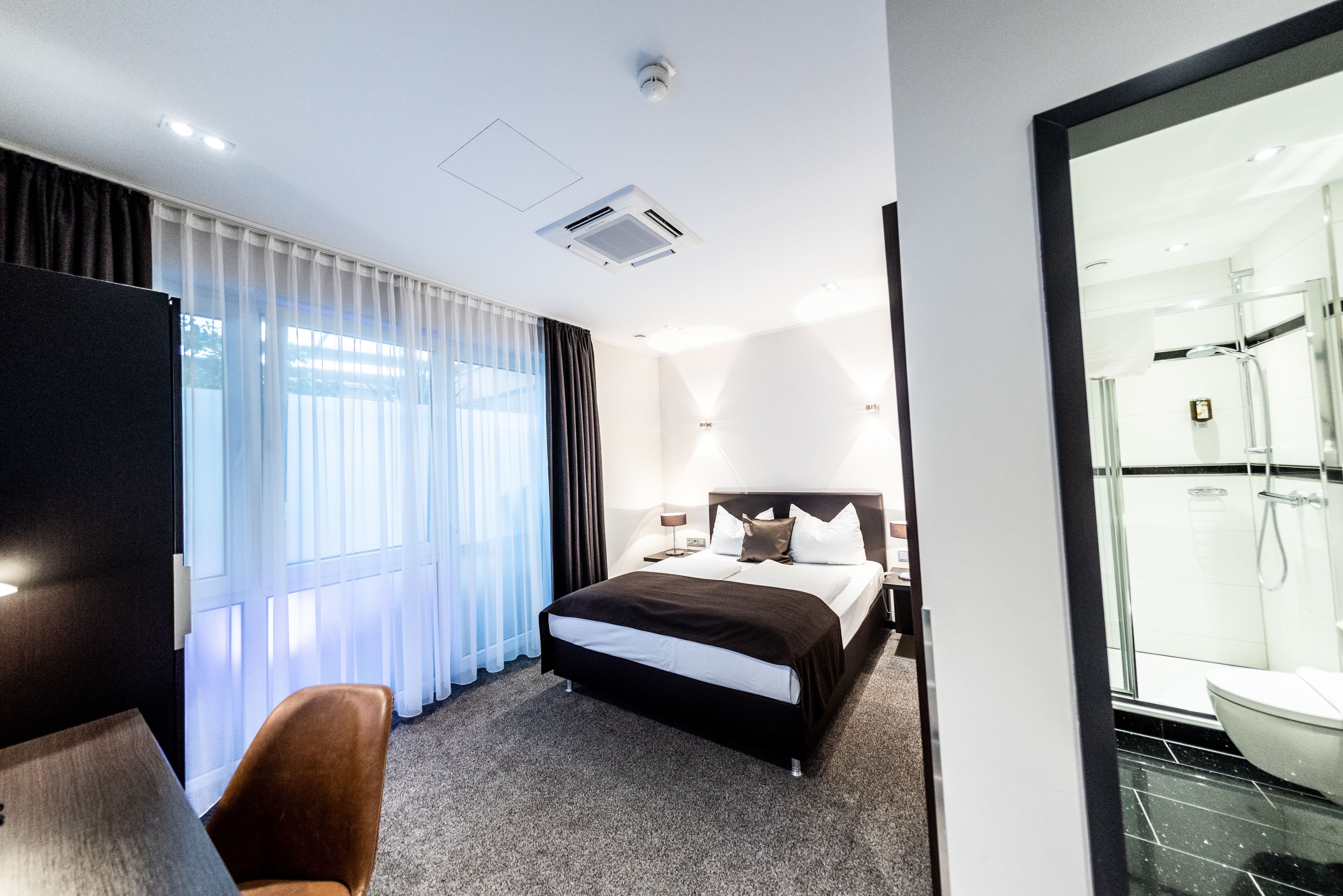 A modern hotel room with a large bed, a desk, a chair, and a glass shower area.