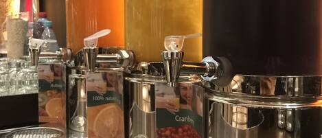Un bar de zumos con varios dispensadores, incluyendo sabores de naranja y arándano, y un gran recipiente con líquido oscuro.