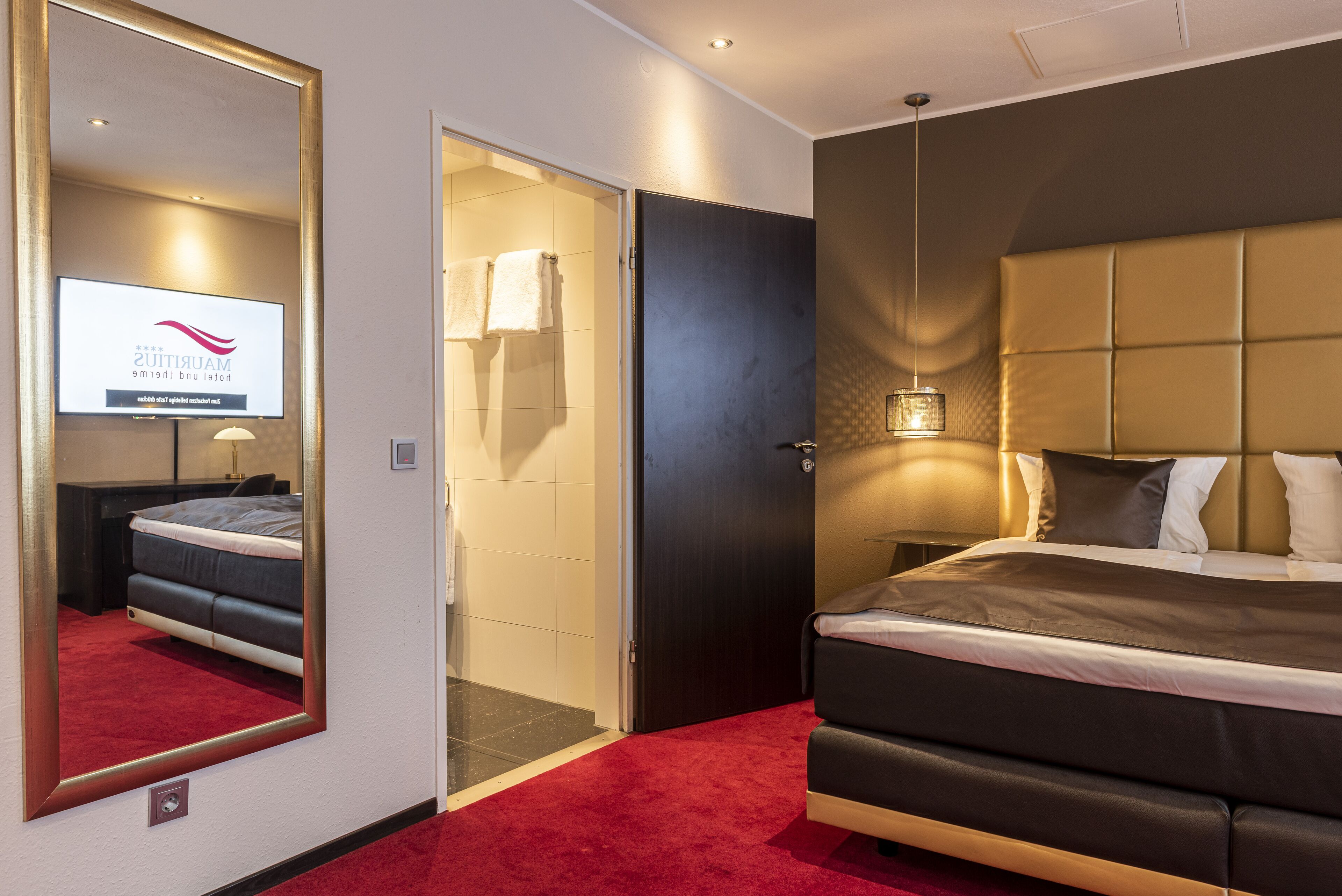 A hotel room with a large bed, a red carpet, a mirror, a television displaying a logo, and a door leading to a bathroom.