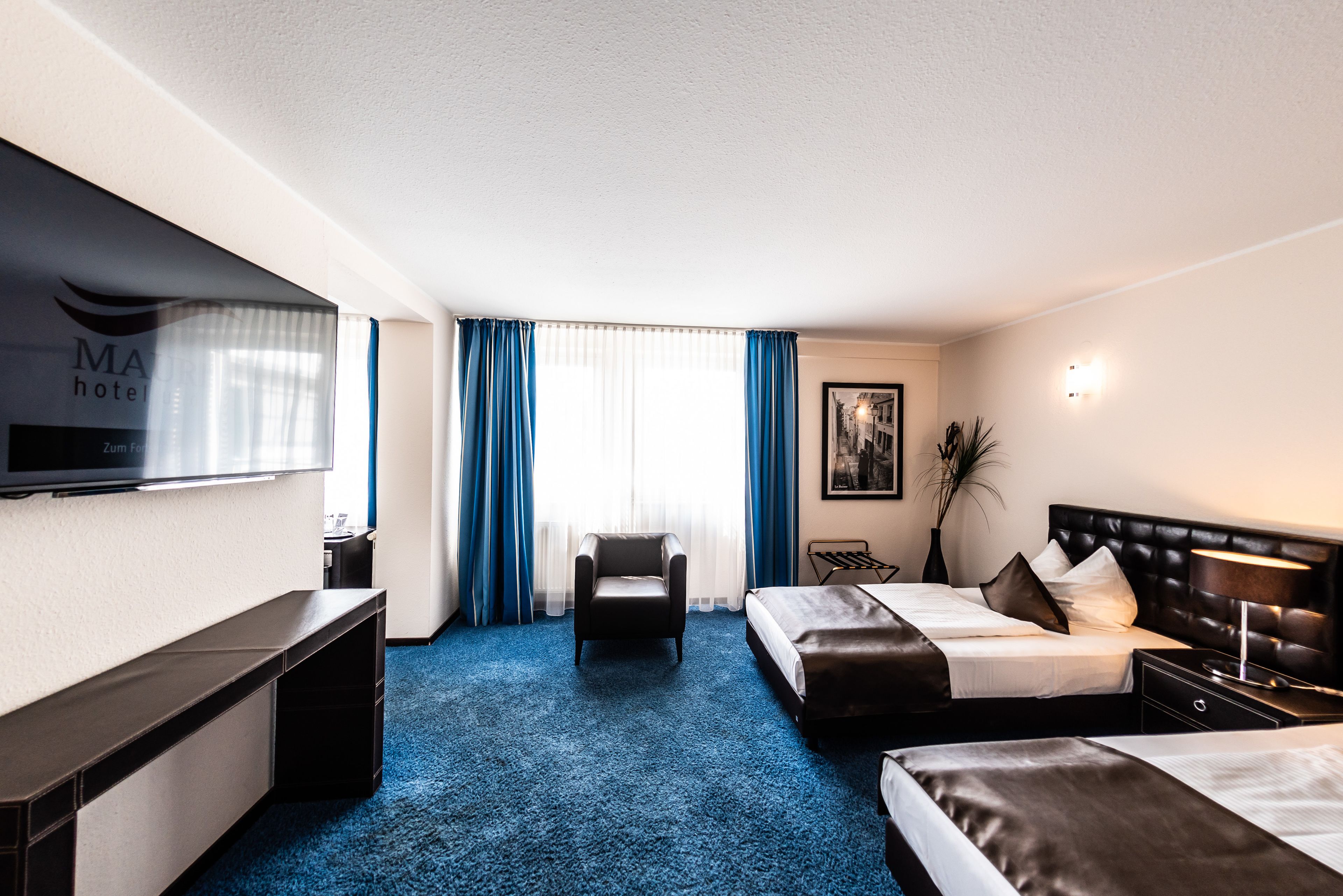 A hotel room with a flat-screen TV, two beds, a chair, a desk, and a large window with curtains.