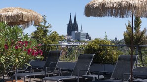 A rooftop terrace with lounge chairs and thatched umbrellas, overlooking a cathedral with twin spires. - Mauritius Hotel & Therme (Cologne)