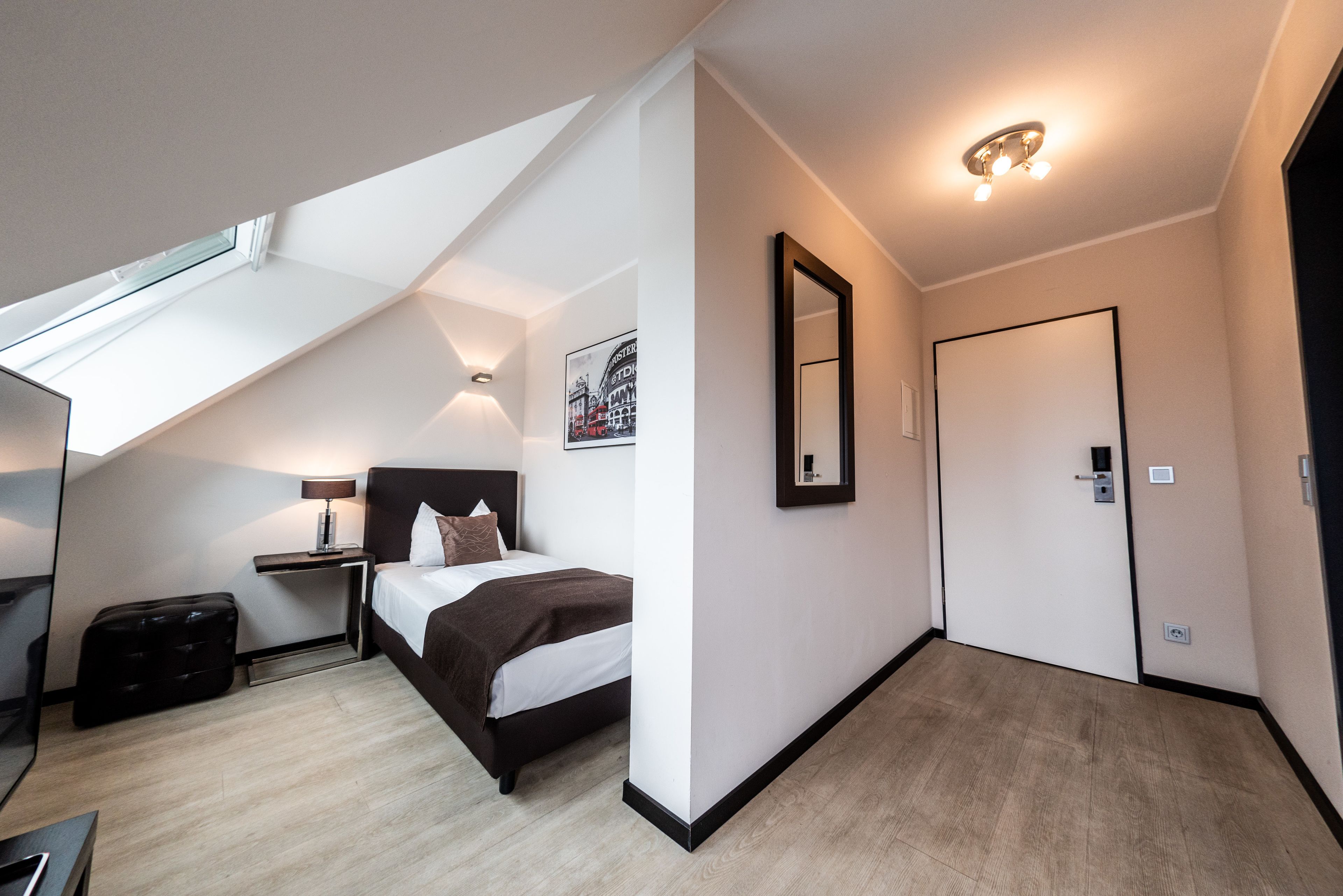 Ein modernes Hotelzimmer mit Dachfenster, einem Bett mit braunem Kopfteil, einem Nachttisch mit Lampe, einem großen Spiegel und einer Tür.