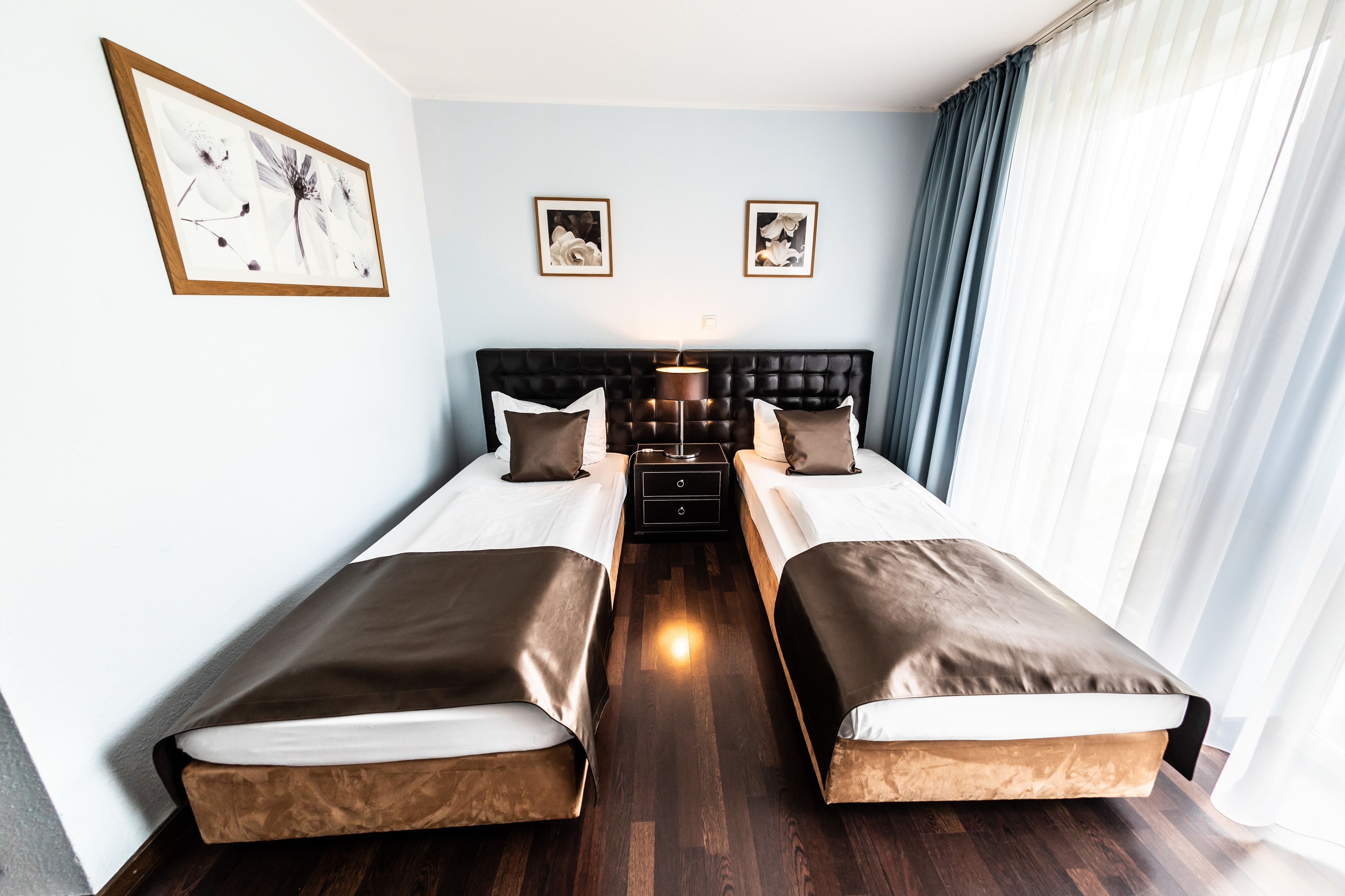 A hotel room with two single beds, a dark headboard, a bedside table, and a large window with curtains.