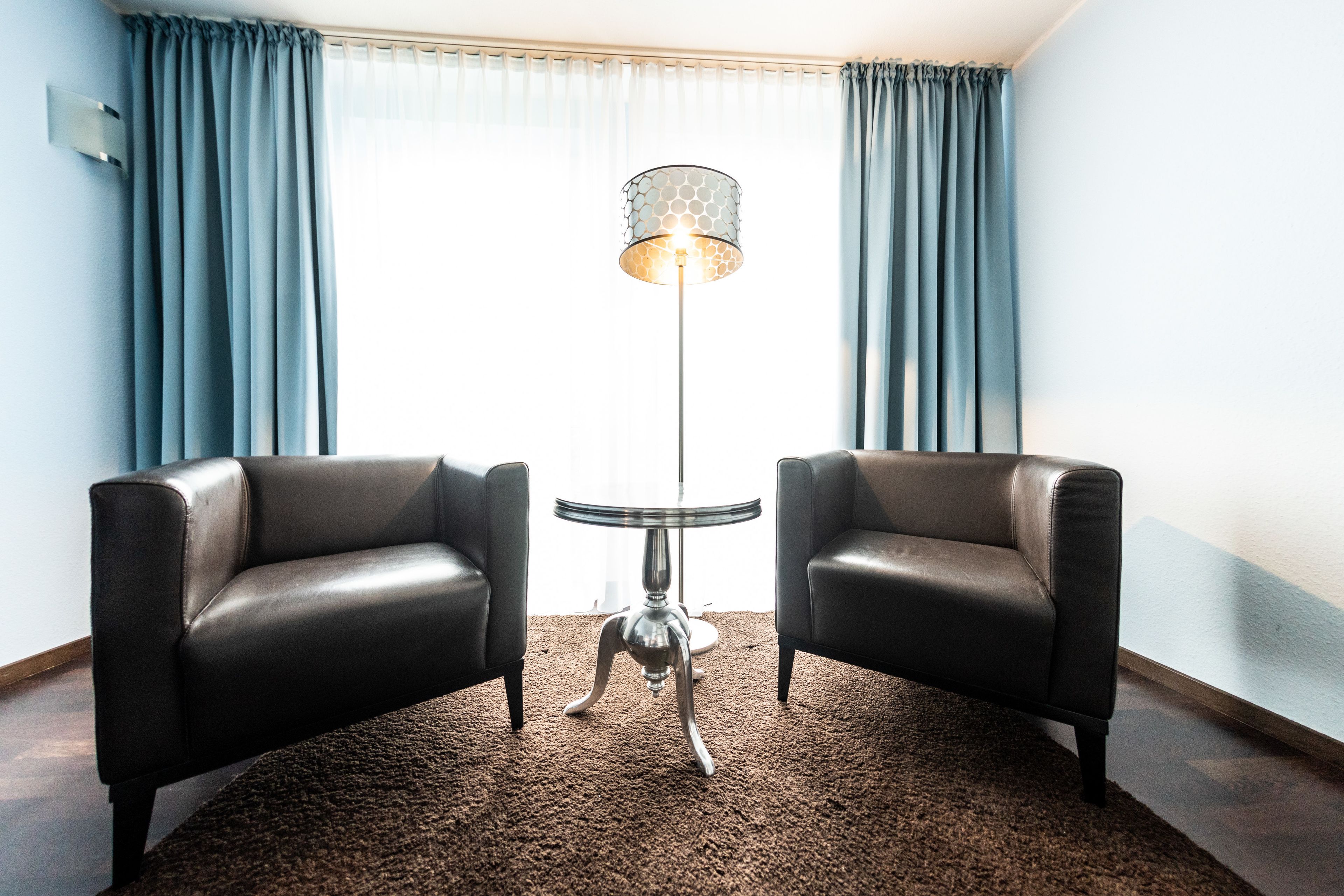 A room with two leather armchairs, a small round table, a floor lamp, and a large window with curtains.
