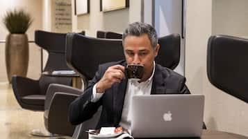 Un hombre con traje sentado a una mesa con una computadora portátil y una taza, en un área de espera moderna con sillones de cuero y una planta en maceta.