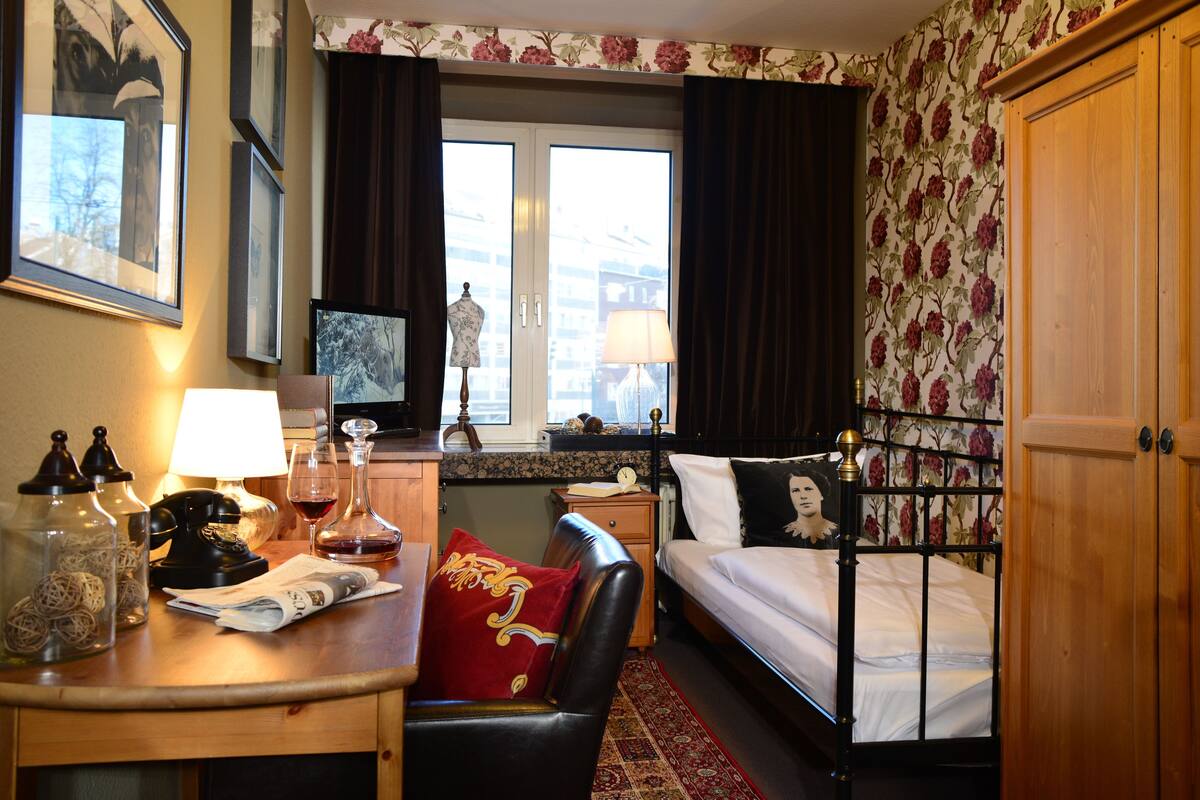 Una habitación de hotel con cama, escritorio con lámpara, silla, armario y ventana con cortinas.