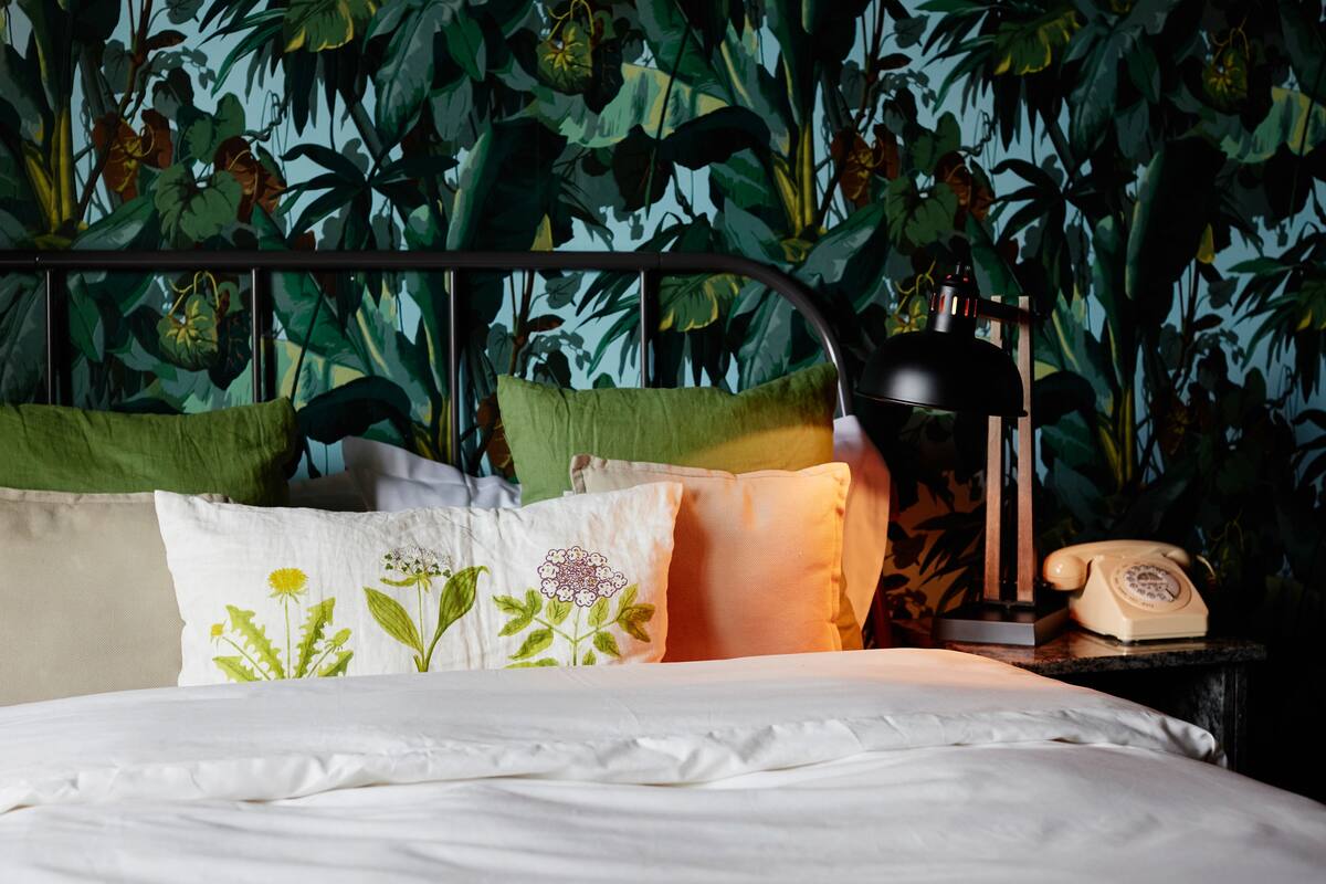 Una cama con ropa blanca, almohadas verdes y beige, y un teléfono giratorio antiguo sobre una mesita de noche.