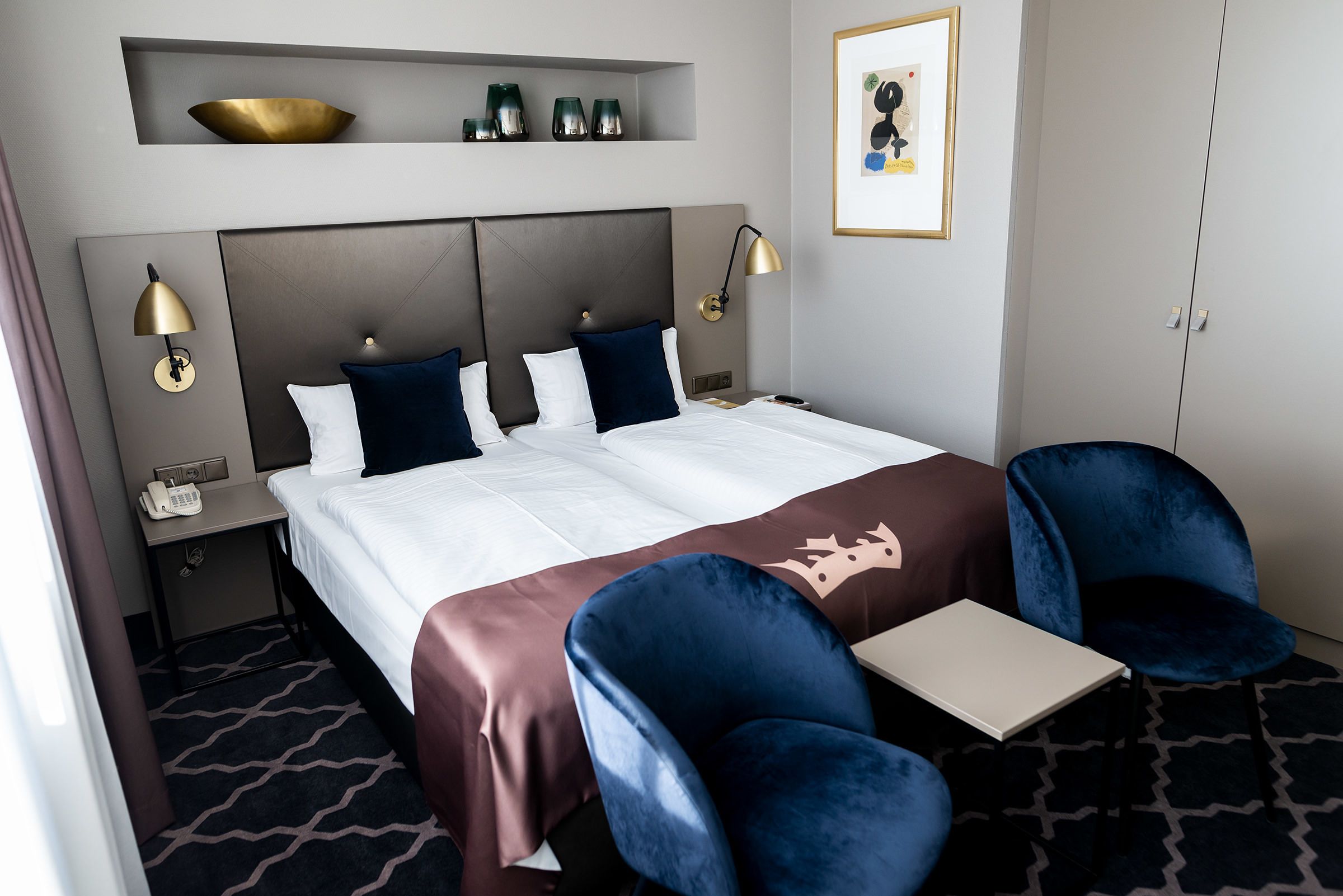 A hotel room with a large bed, two bedside lamps, a desk, two blue chairs, a small table, a phone on the nightstand, and a framed picture on the wall.